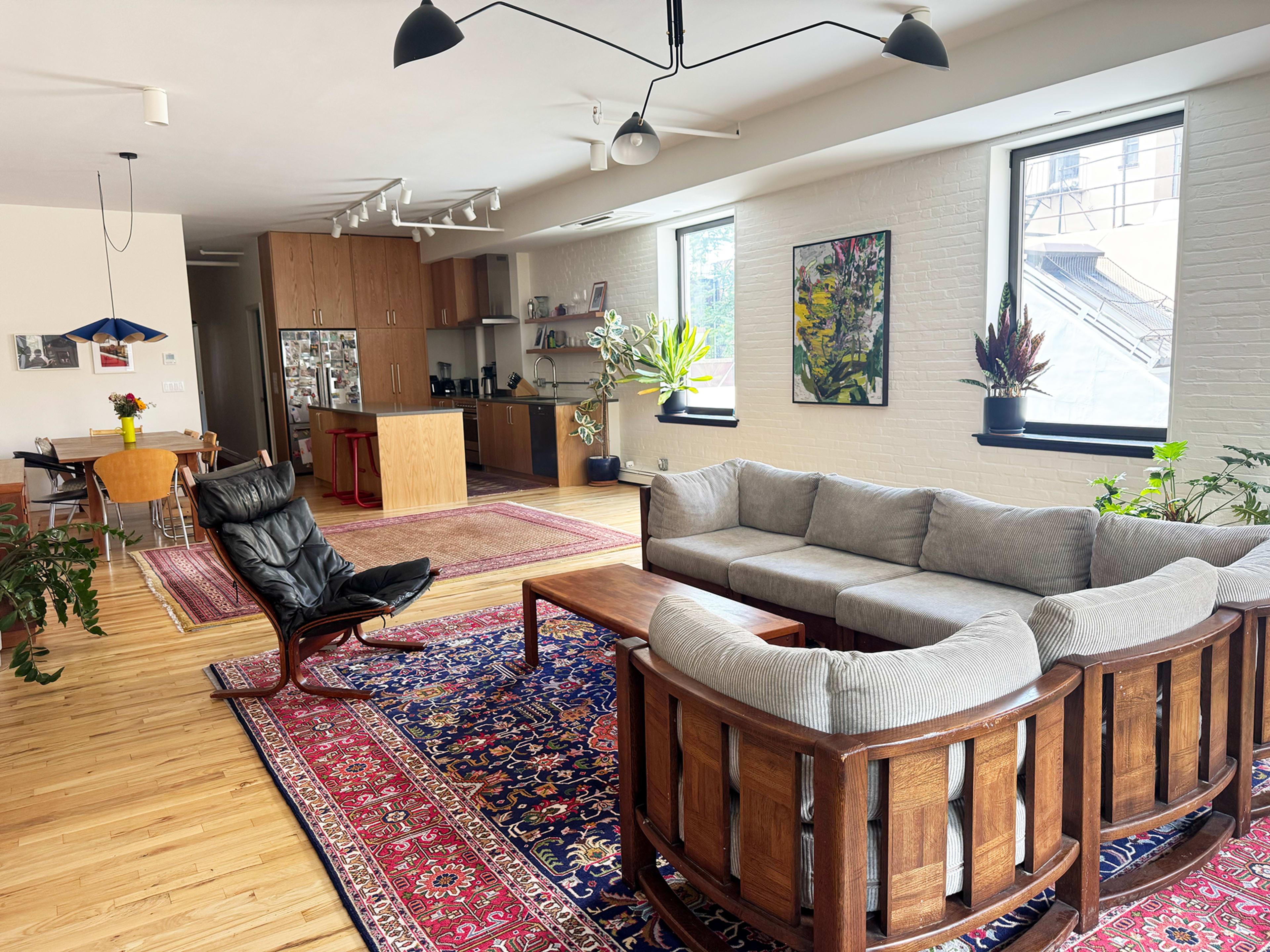 The image shows a modern, open-concept living space with a gray sectional sofa, wooden furniture, and large windows allowing natural light, alongside a dining area and a kitchen in the background.