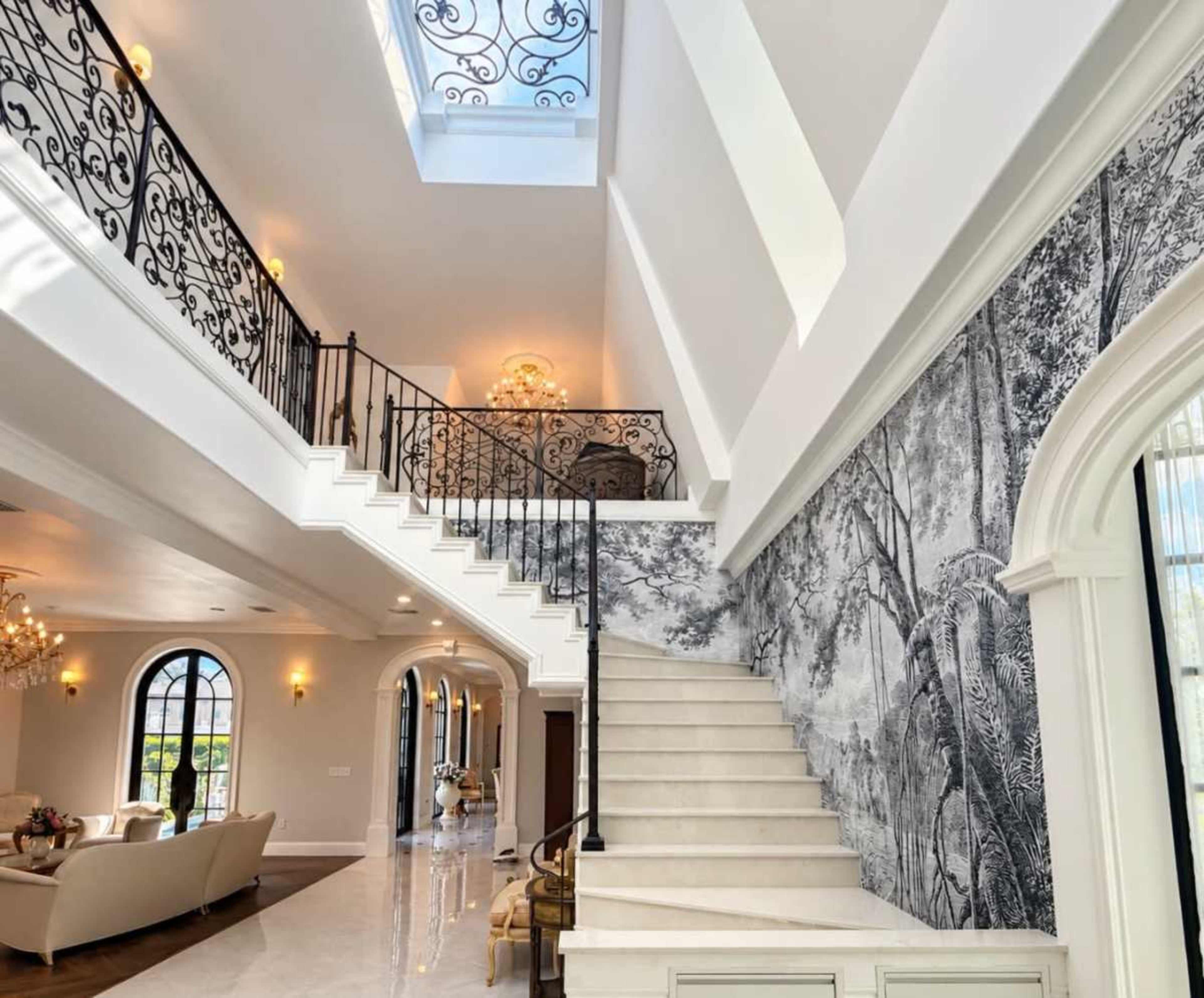 The image shows a spacious entryway with a grand staircase featuring ornate wrought-iron railing, illuminated by natural light from a skylight above.