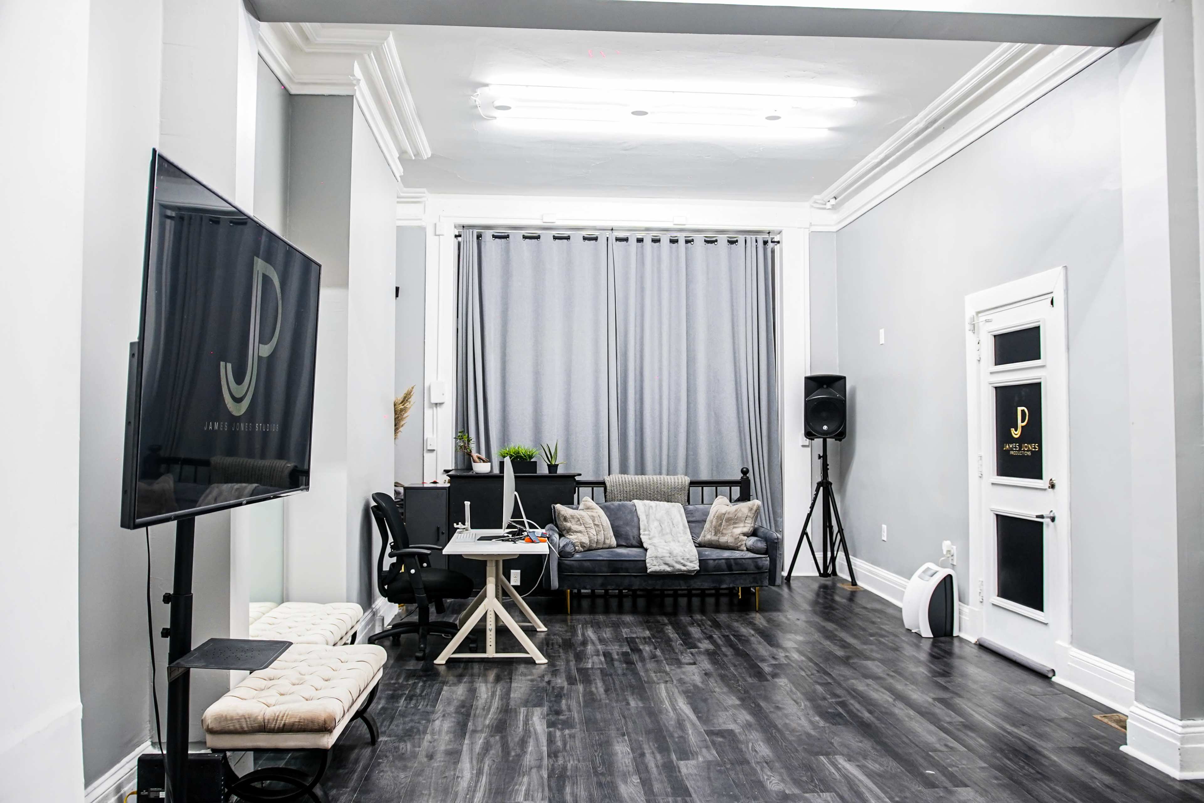 A modern, minimalist office or studio space with a dark wooden floor, a comfortable couch, a desk with a computer, and gray curtains covering a window.