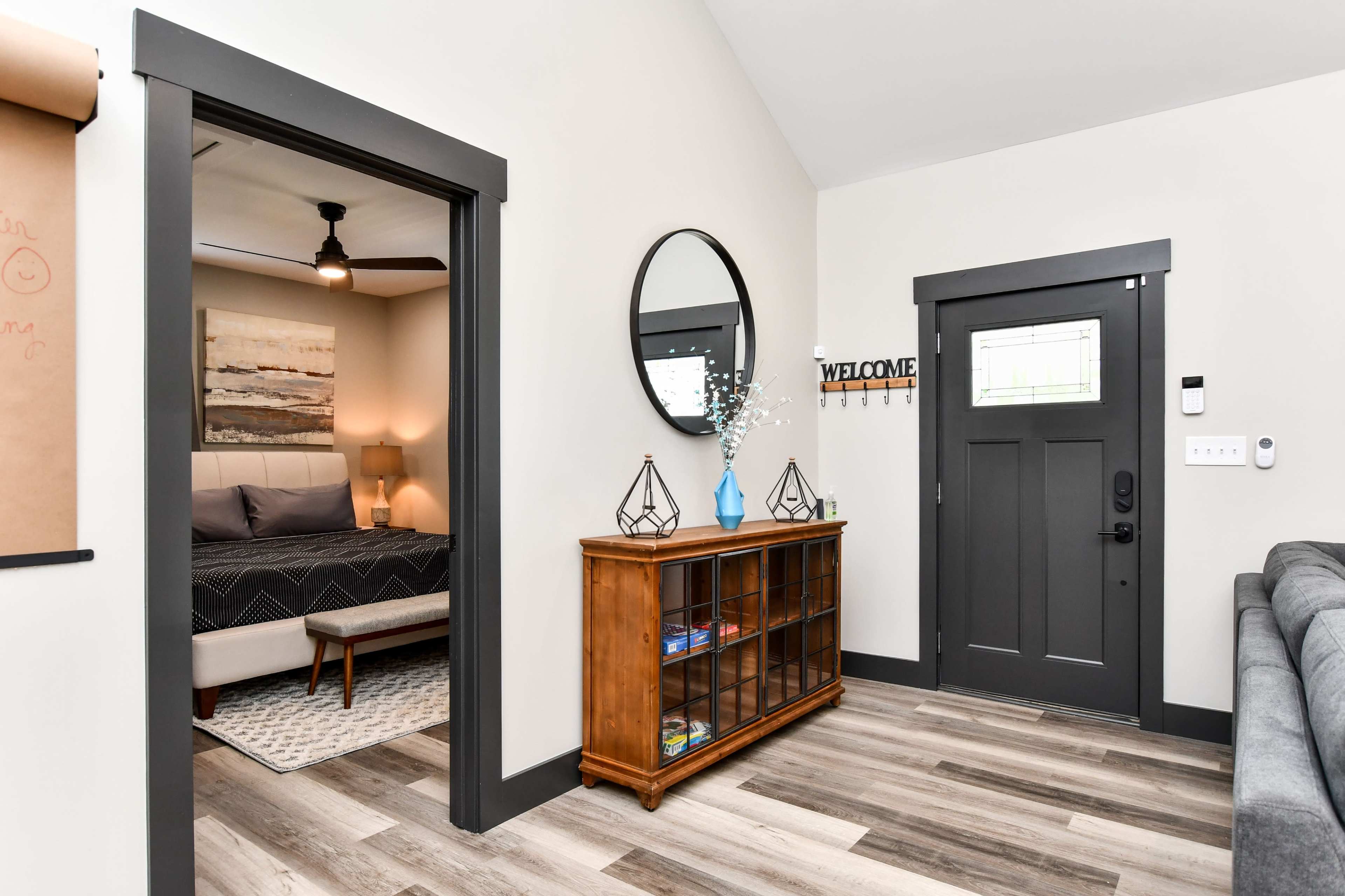 A cozy entryway featuring a storage cabinet, a circular mirror, and a door leading to a bedroom, with a welcoming ambiance created by natural light and neutral tones.