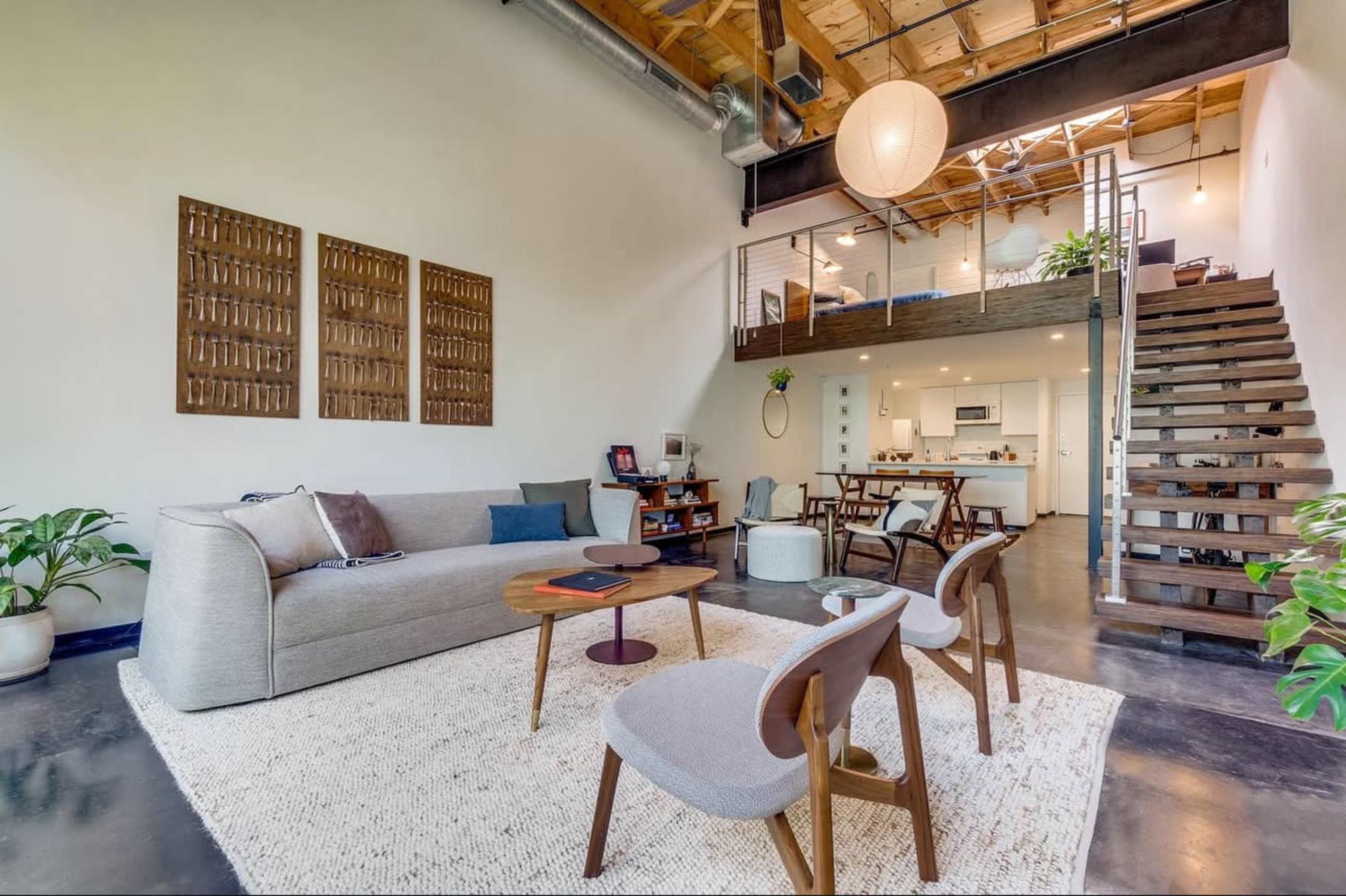 The image shows a modern, spacious living area with a sofa, a coffee table, and two chairs, featuring a staircase that leads to an upper level with a dining area and kitchen.