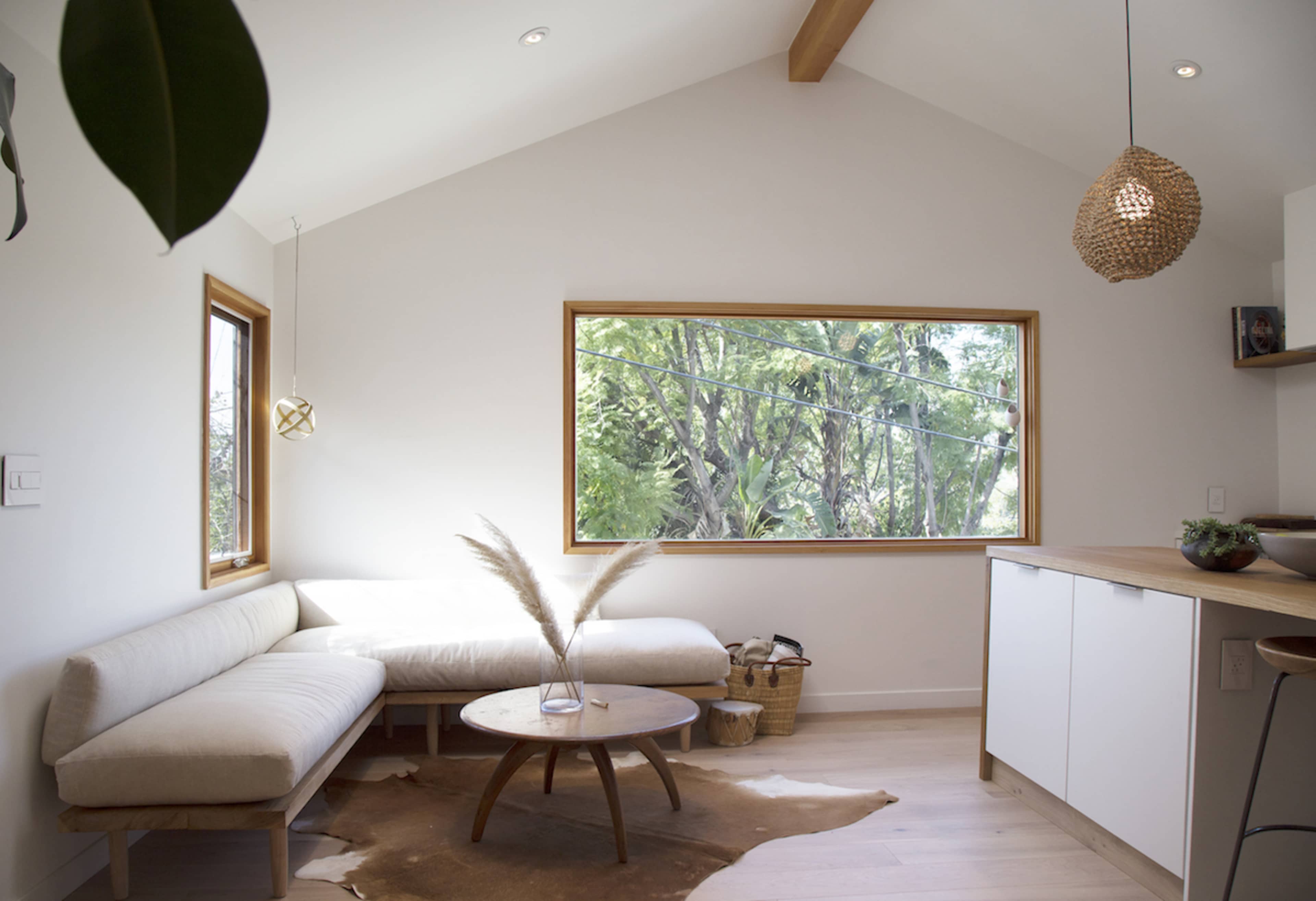 A brightly lit, modern living space featuring a cozy white couch, a round wooden table atop a decorative rug, and large windows overlooking lush greenery.