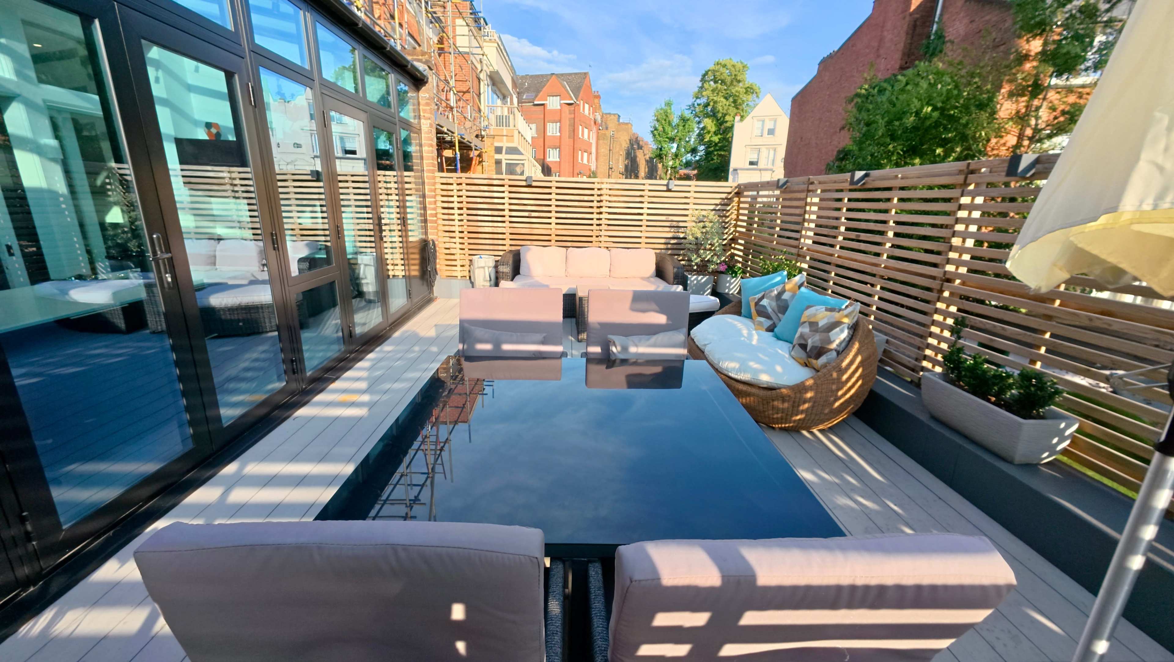 A modern outdoor terrace features a glass table surrounded by several couches and seating areas, framed by wooden lattice partitions and greenery.