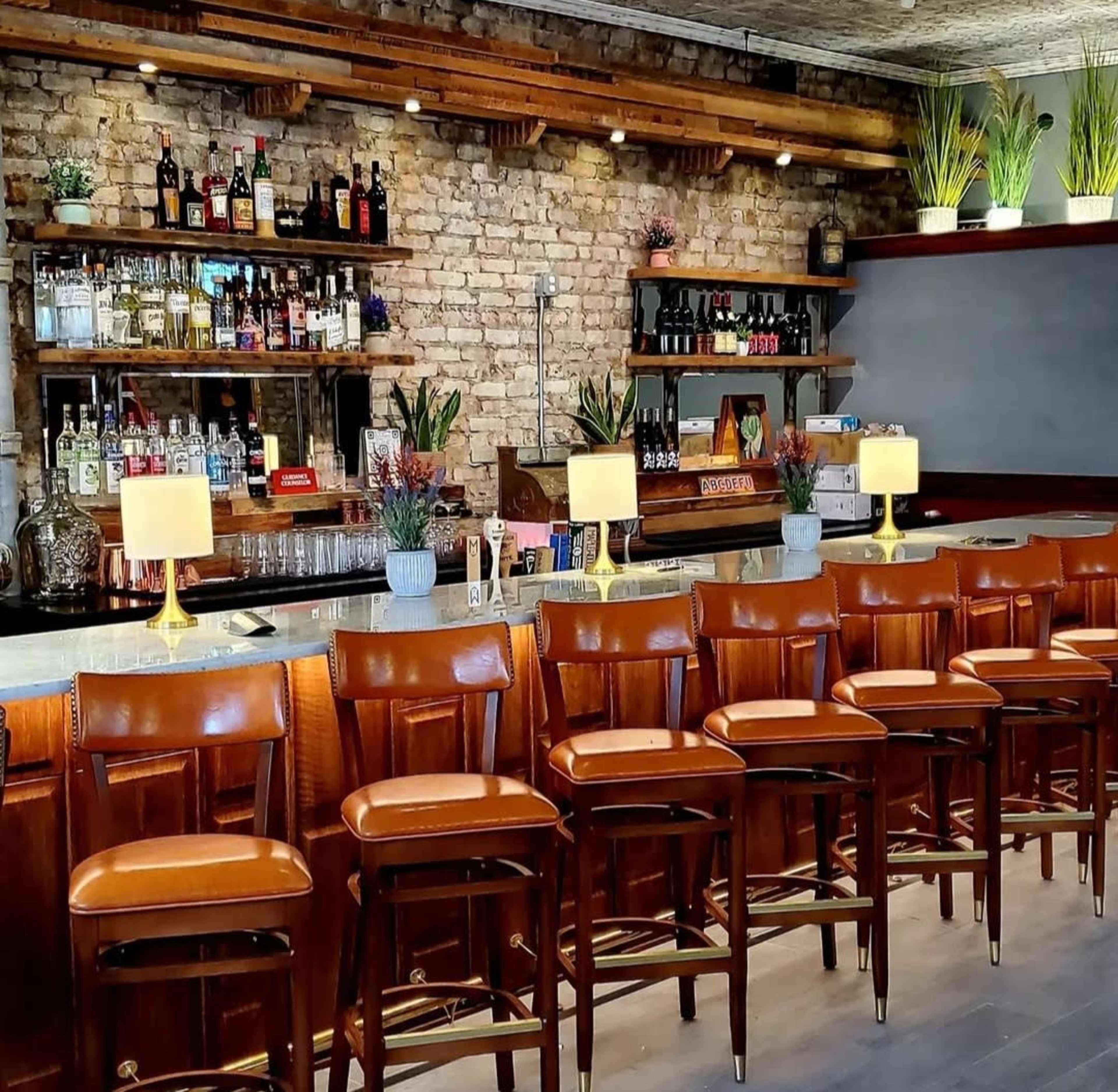 The image shows a bar area with a wooden counter and several high-backed chairs, against a backdrop of exposed brick and shelves filled with liquor bottles.