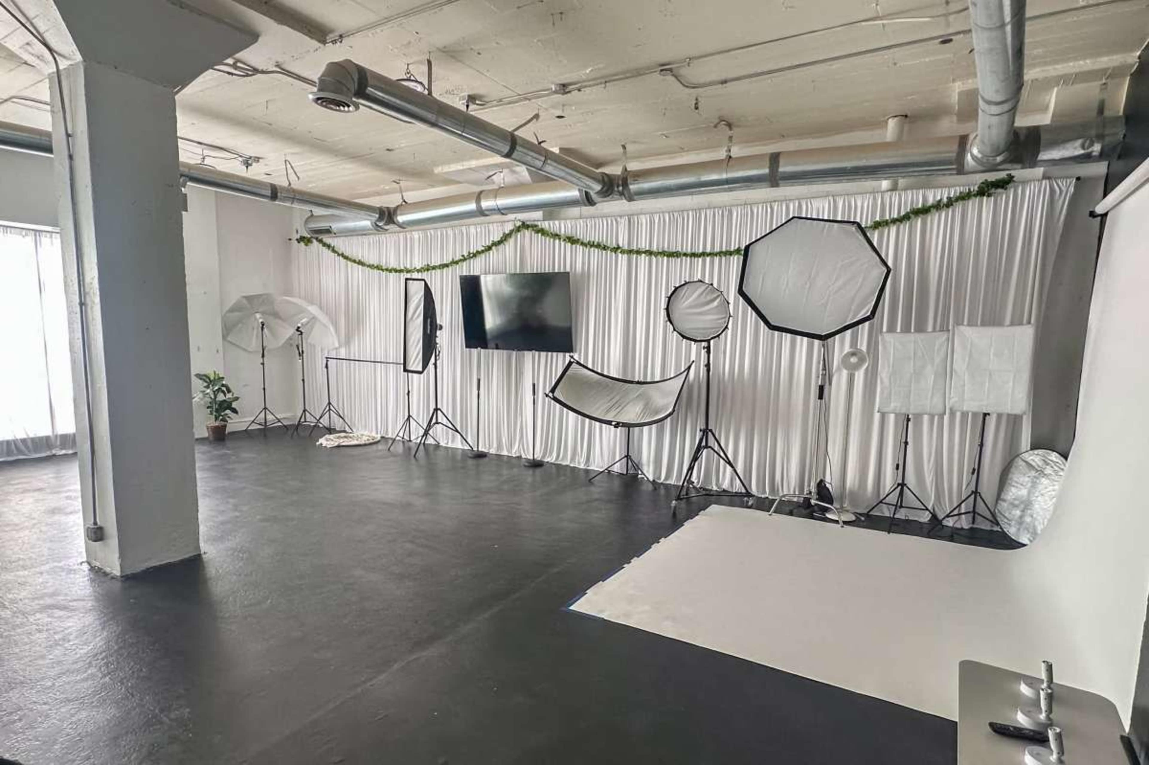 The image shows a photography studio featuring various lighting equipment, backdrops, and a large screen set against white drapes.