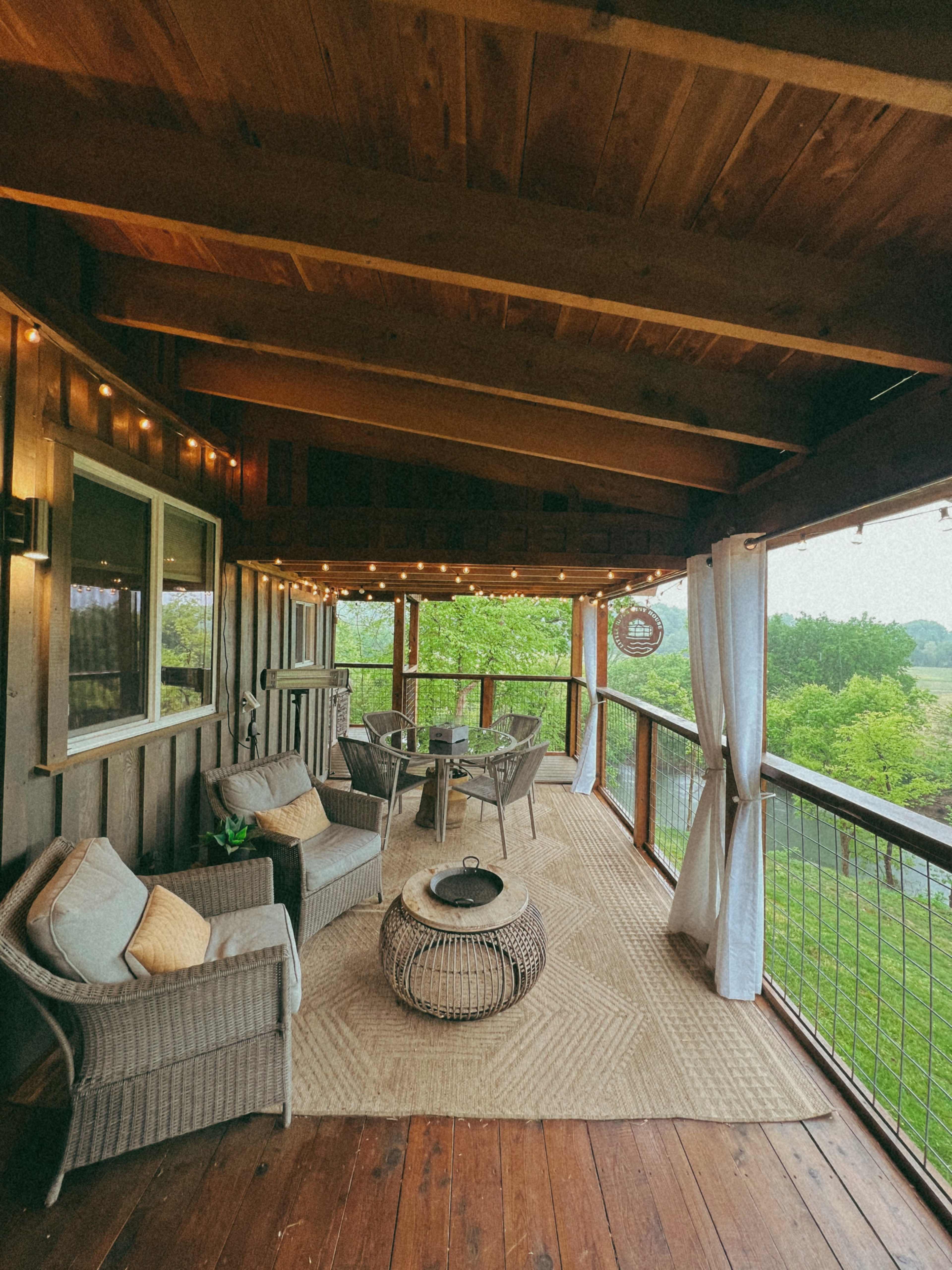 A covered wooden porch features a seating area with wicker furniture, a round table, and decorative string lights, overlooking a scenic view.