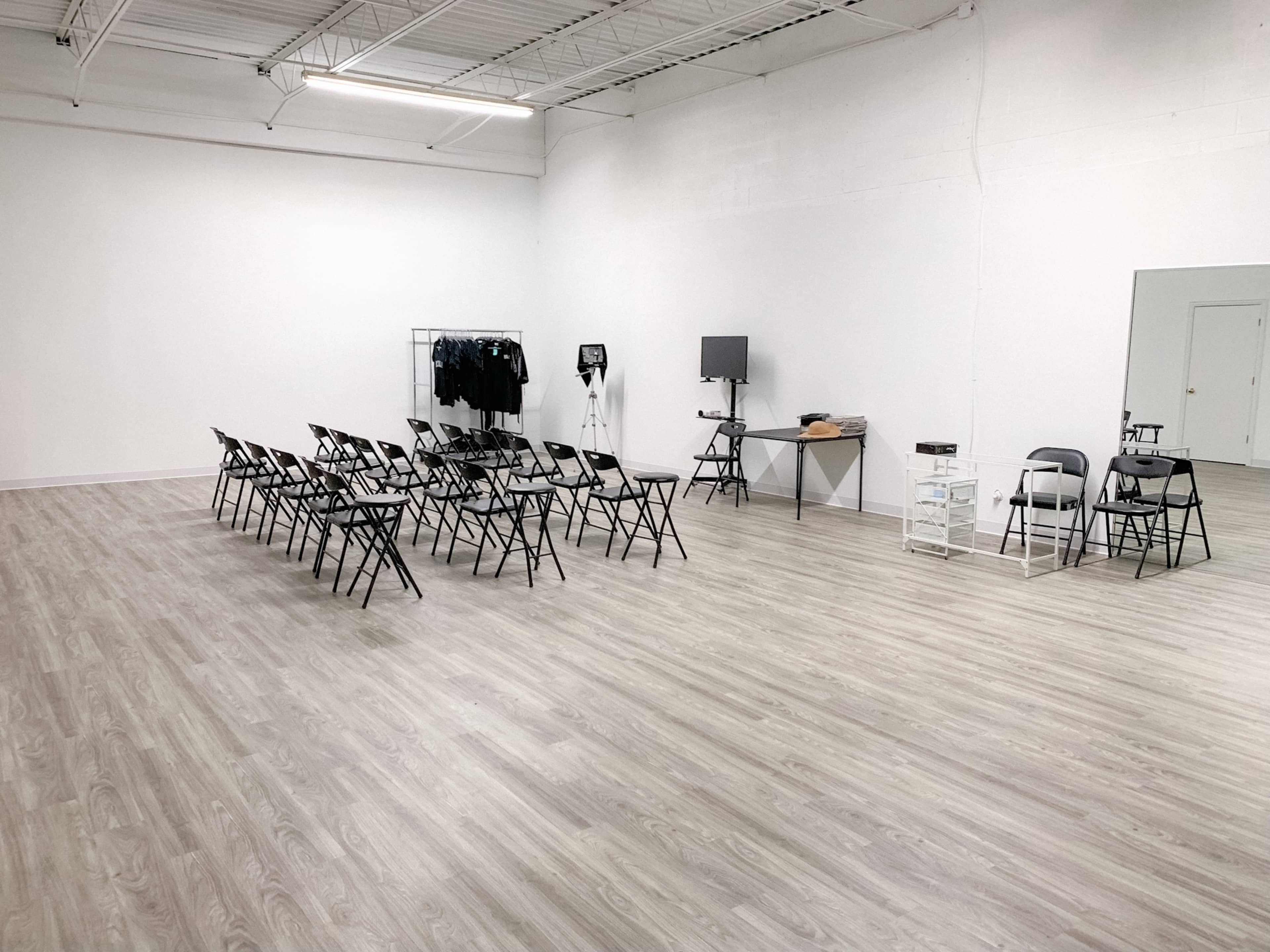 A spacious, empty room with a row of folding chairs facing a wall, a table with equipment, and a large mirror on the opposite side.