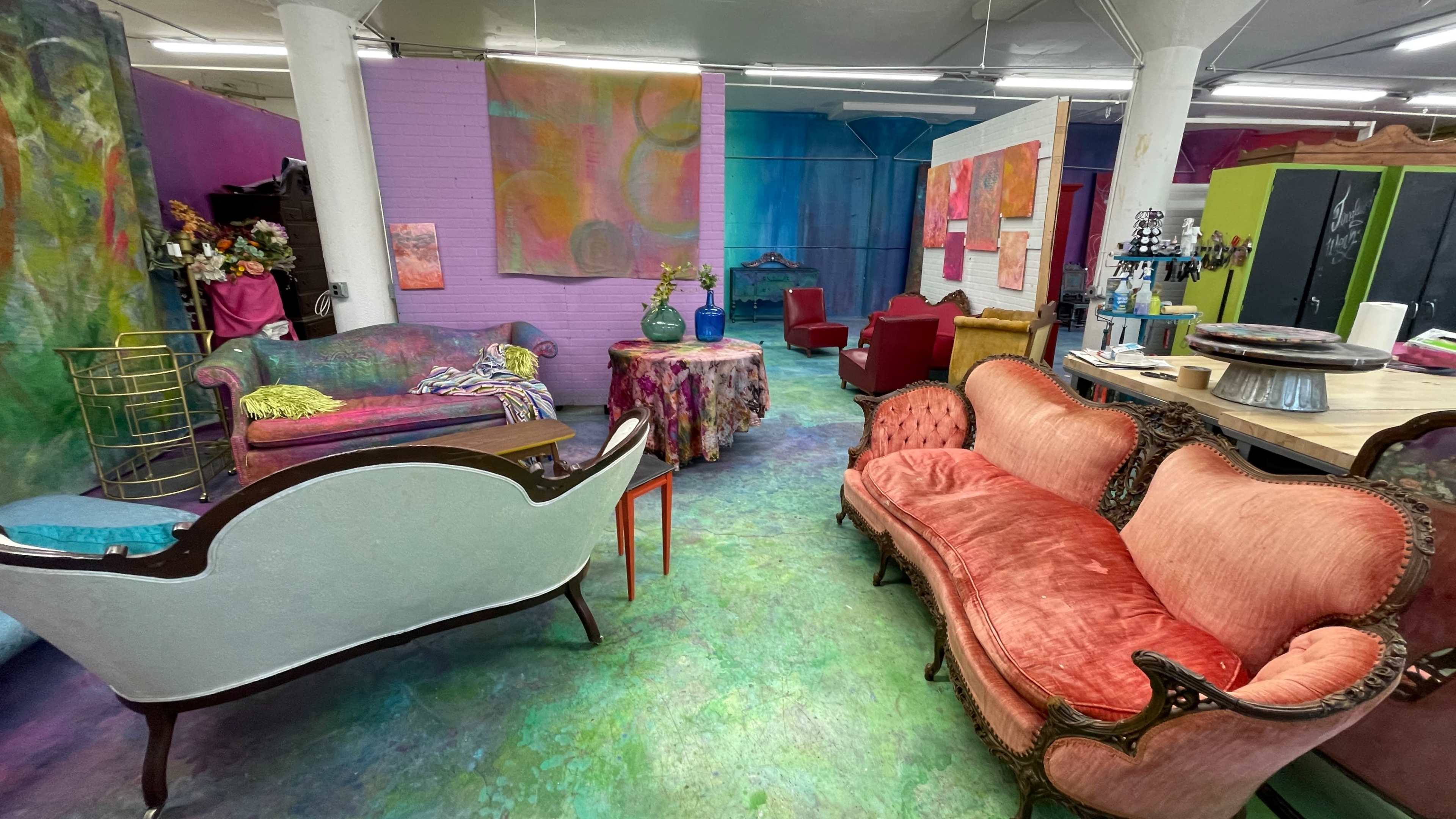 The image depicts a colorful interior space with eclectic furniture, including several vintage sofas and art-covered walls.