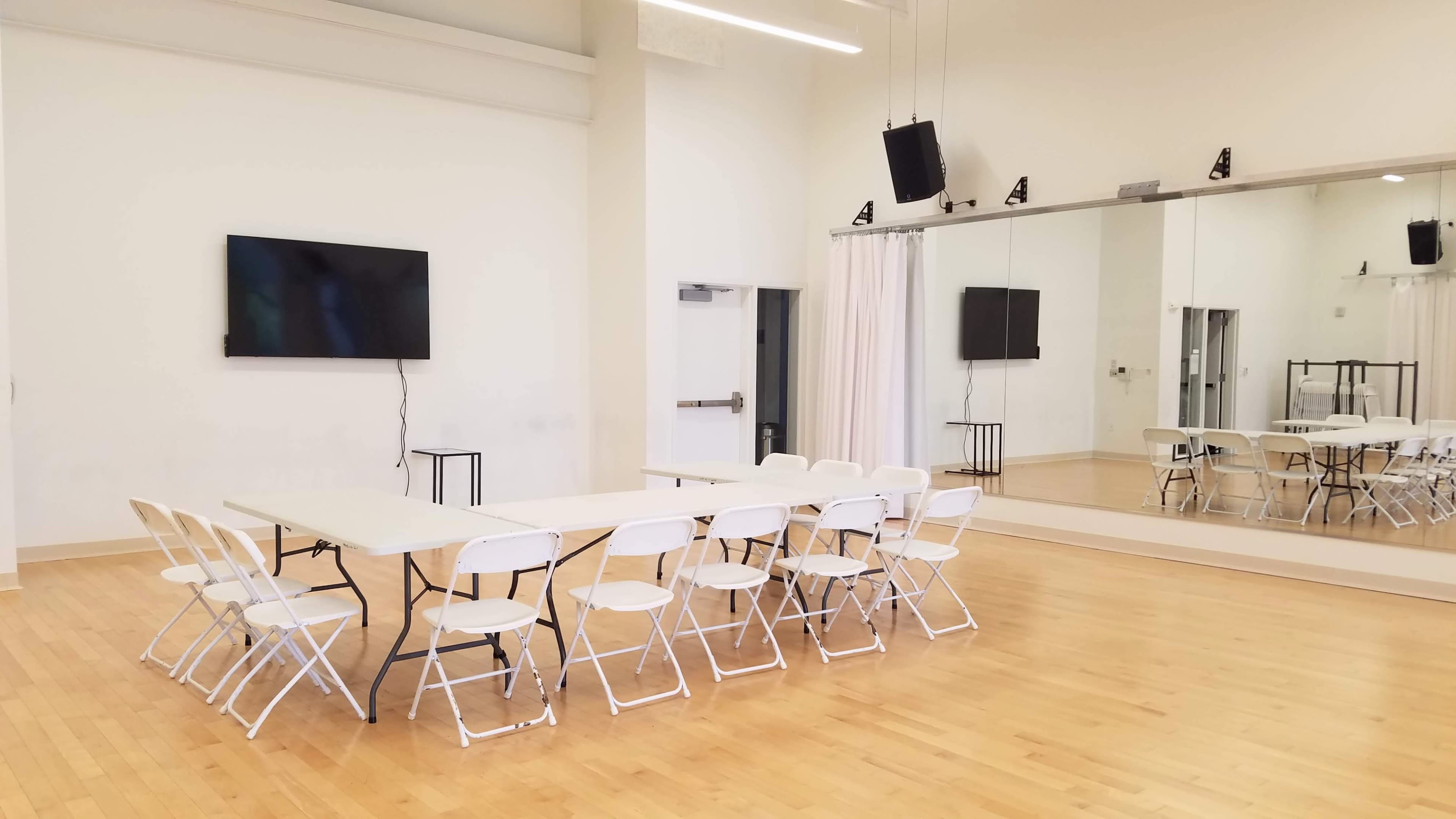 A meeting room features a long table with white folding chairs arranged around it, a large screen on one wall, and a mirrored wall opposite.