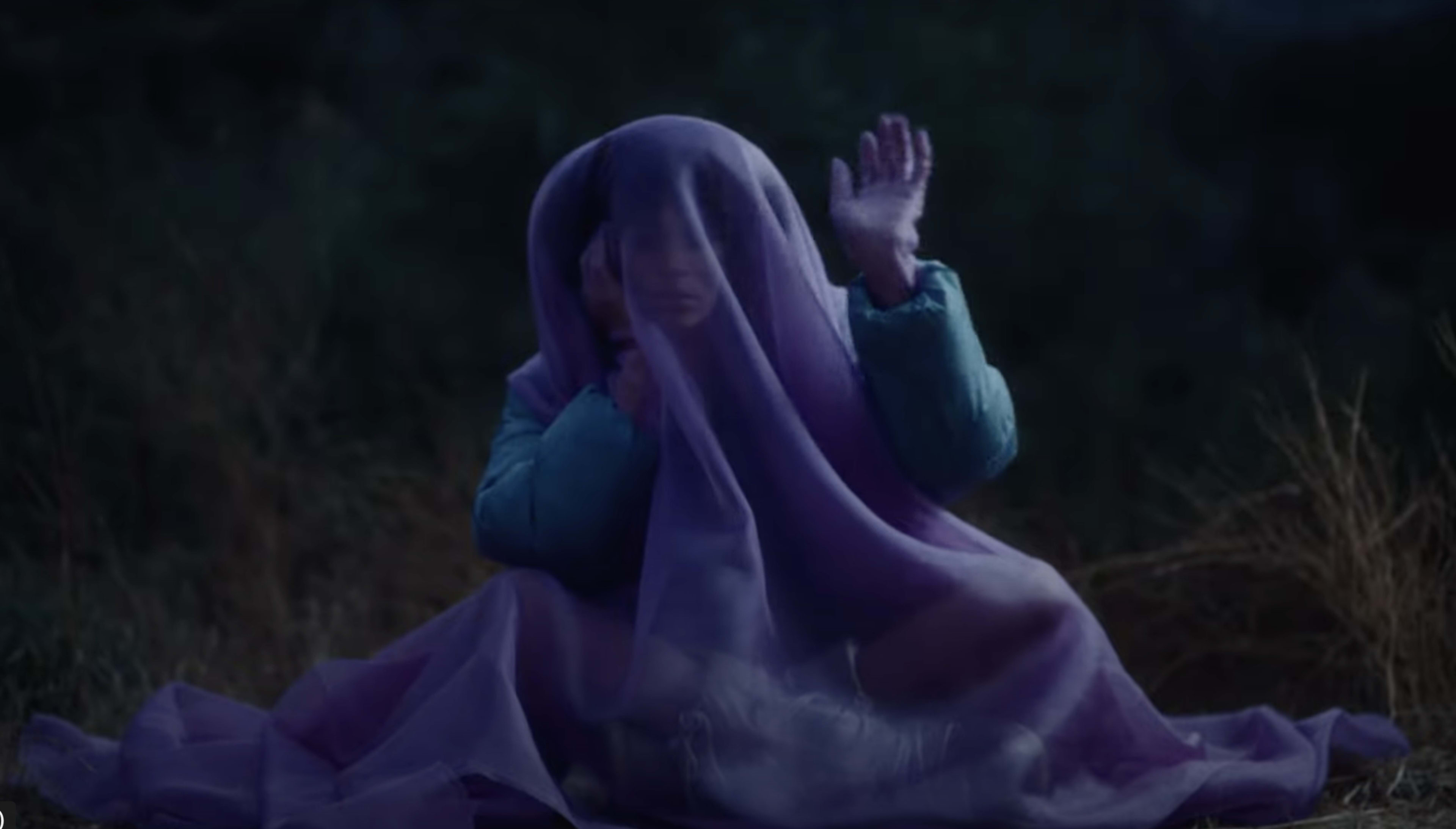 A person is sitting on the ground, partially covered by a translucent purple fabric, with their hand raised in a gesture.