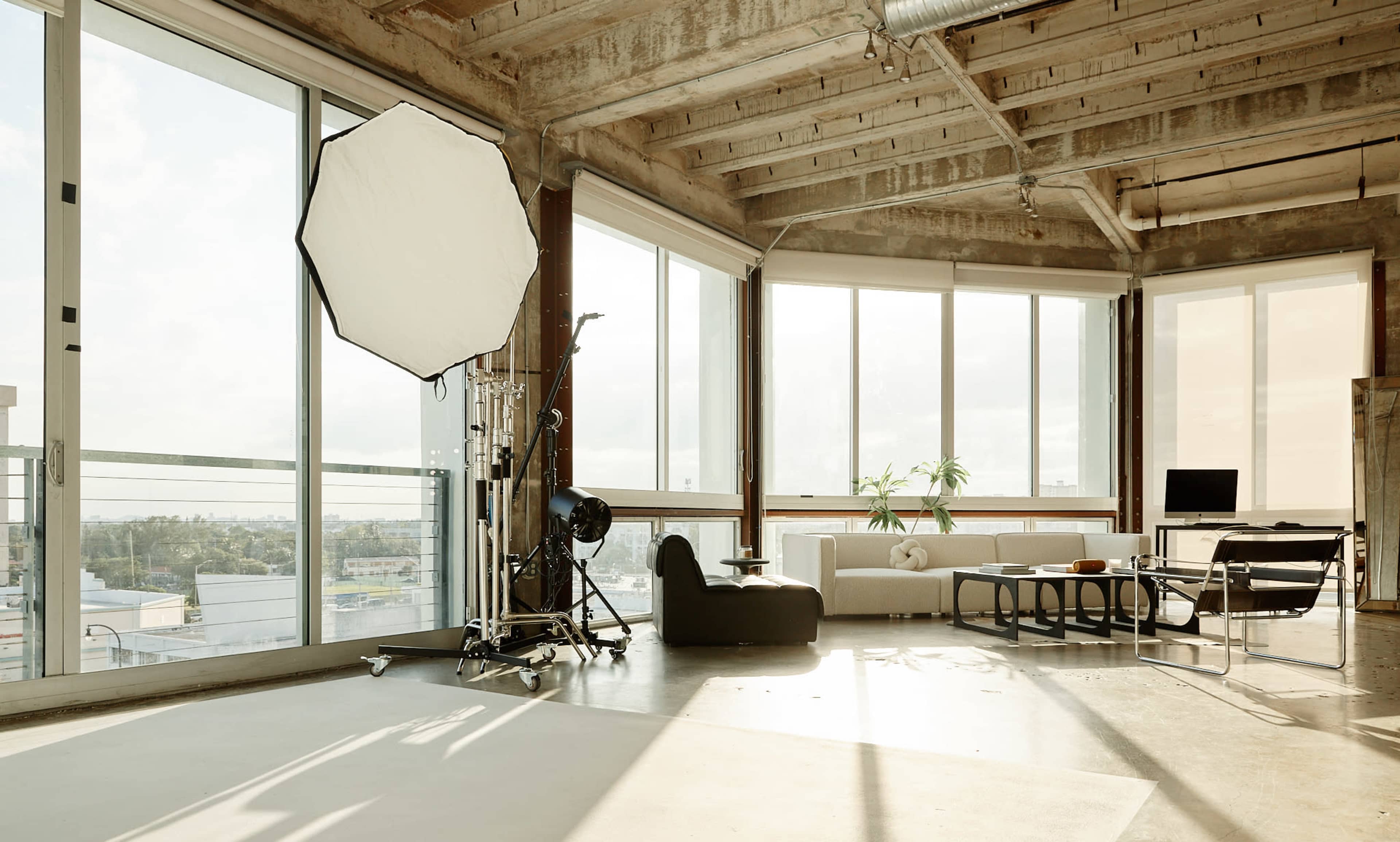 A spacious, industrial-style room features large windows, modern furniture, and equipment for photography.