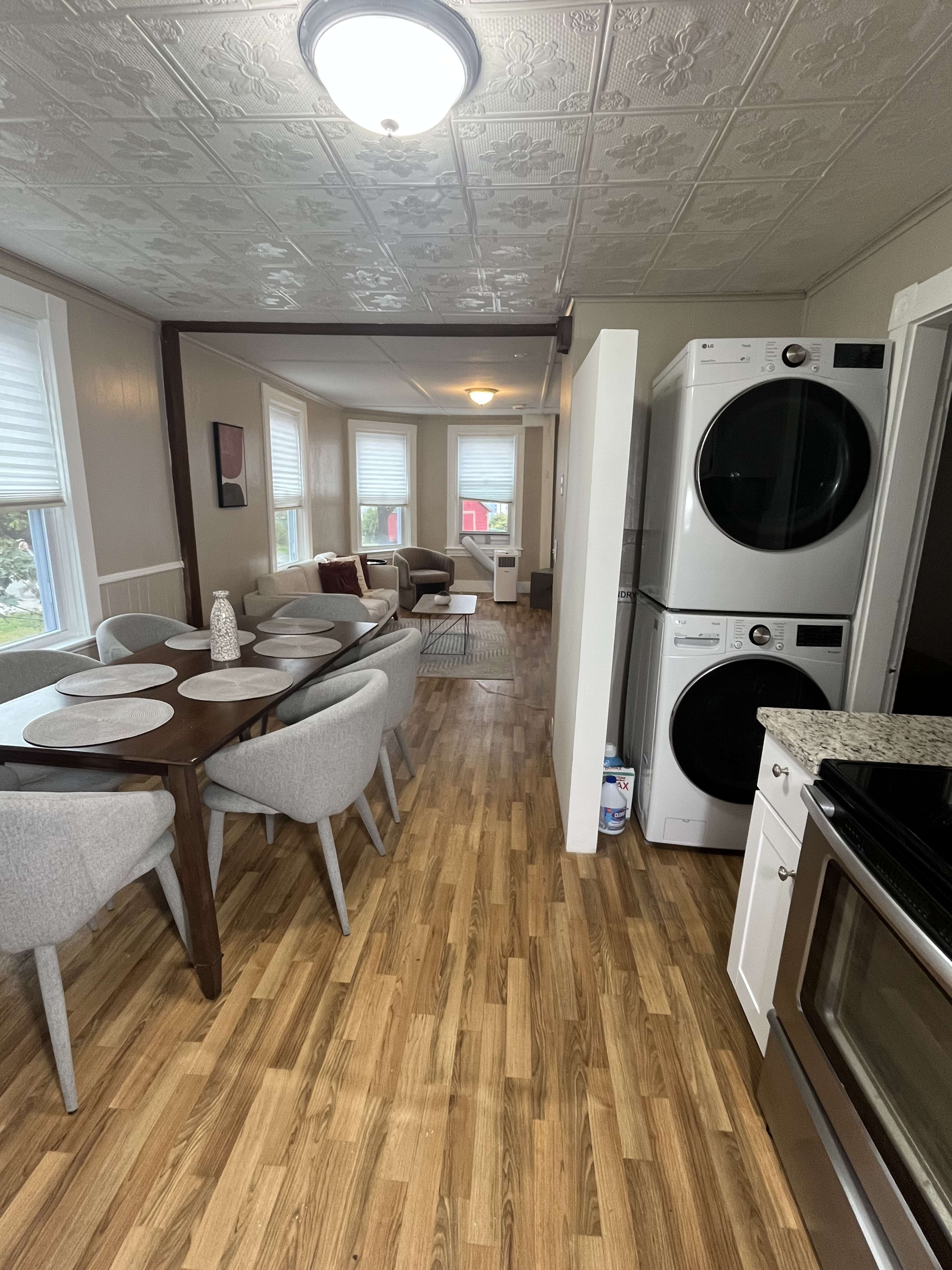 The image shows a modern kitchen and dining area featuring a round dining table with gray chairs, a stacked washer and dryer, and a cozy living space with a sofa and large windows.