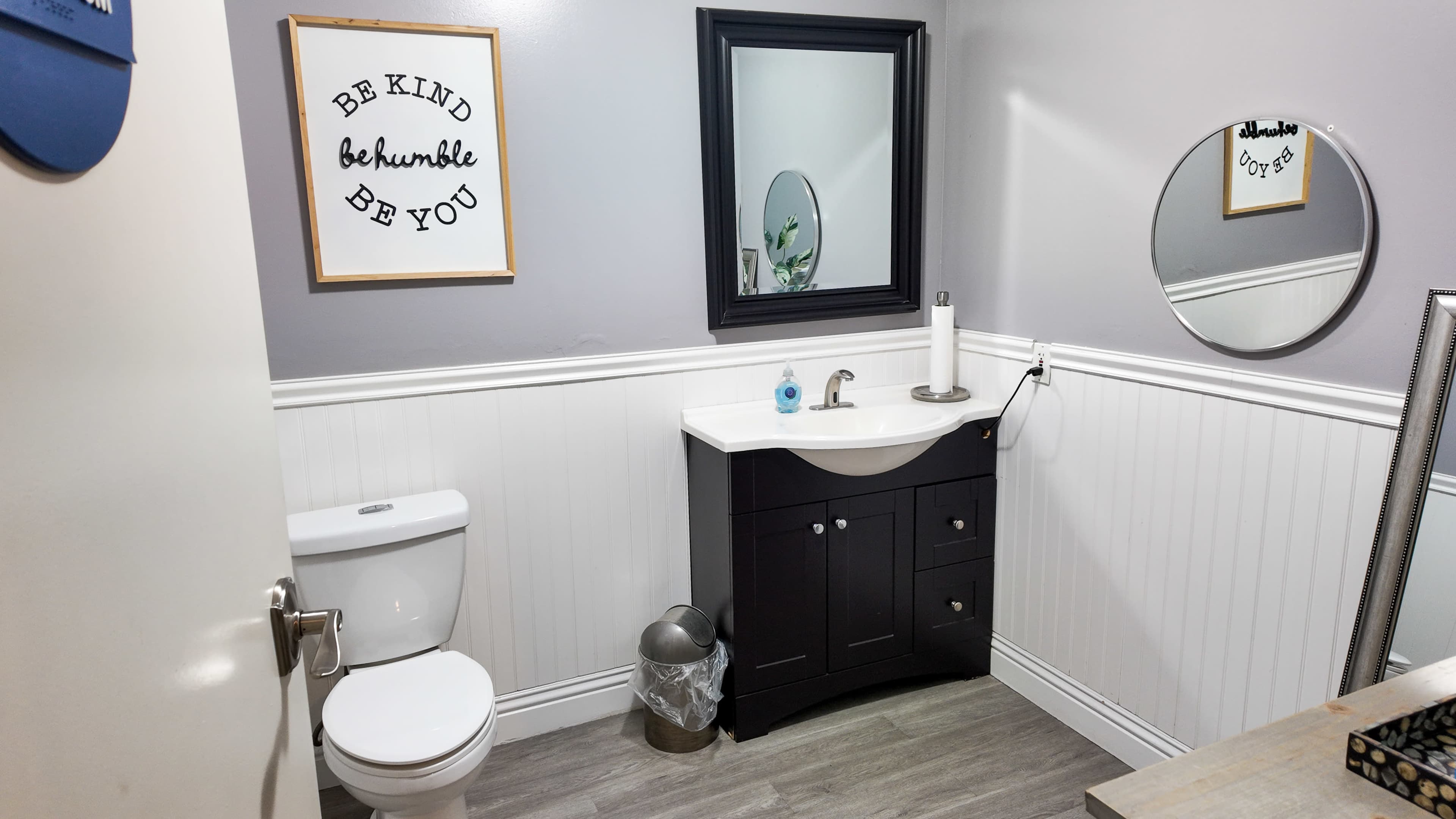 The image shows a bathroom featuring a toilet, a sink with a dark cabinet, and two mirrors mounted on the gray walls, one of which has a decorative sign that reads "BE KIND."