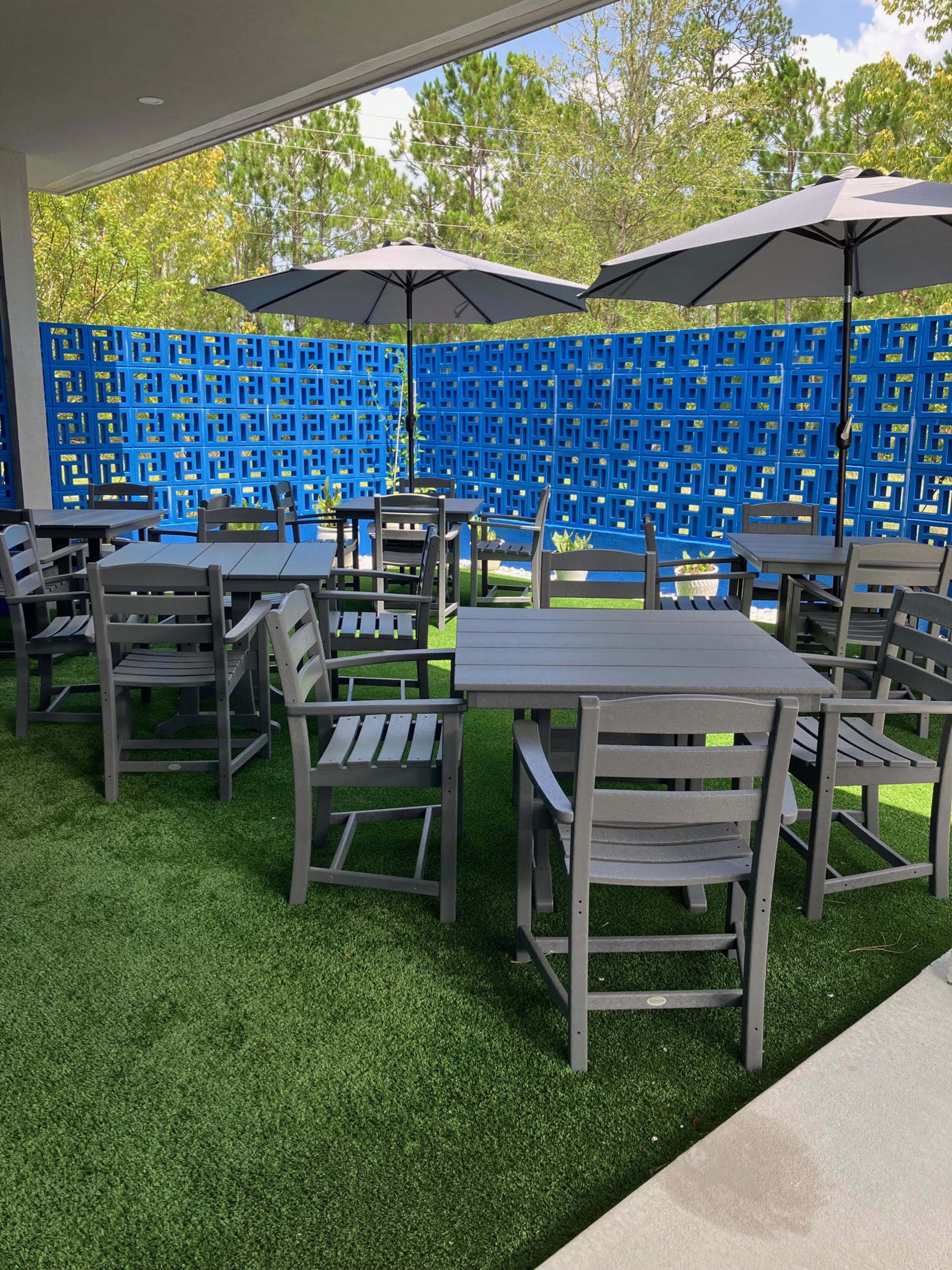 The image shows a patio area with several gray outdoor tables and chairs arranged on artificial grass, shaded by large umbrellas, and a blue patterned wall in the background.