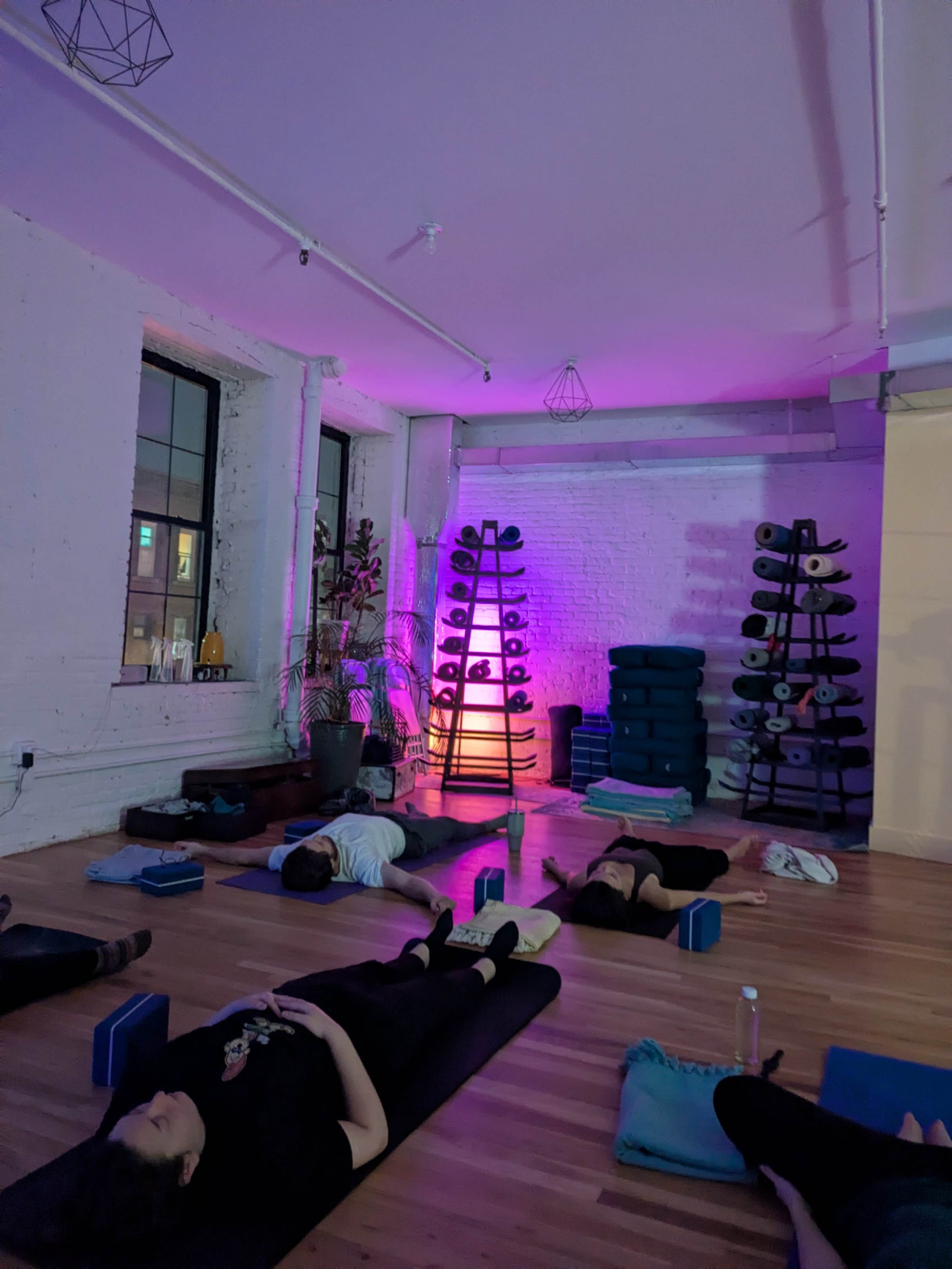 People are practicing yoga or meditation in a dimly lit studio with purple lighting and various yoga props arranged nearby.