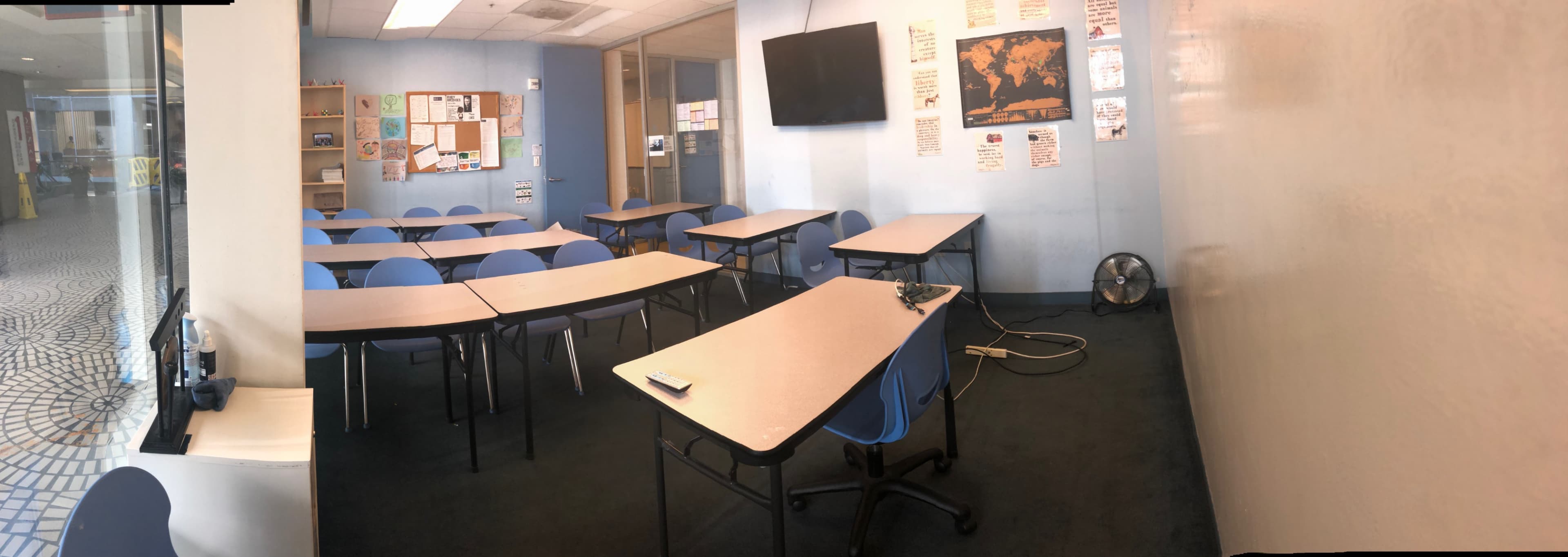A classroom with rows of tables and chairs, a wall-mounted television, and a fan on the floor.