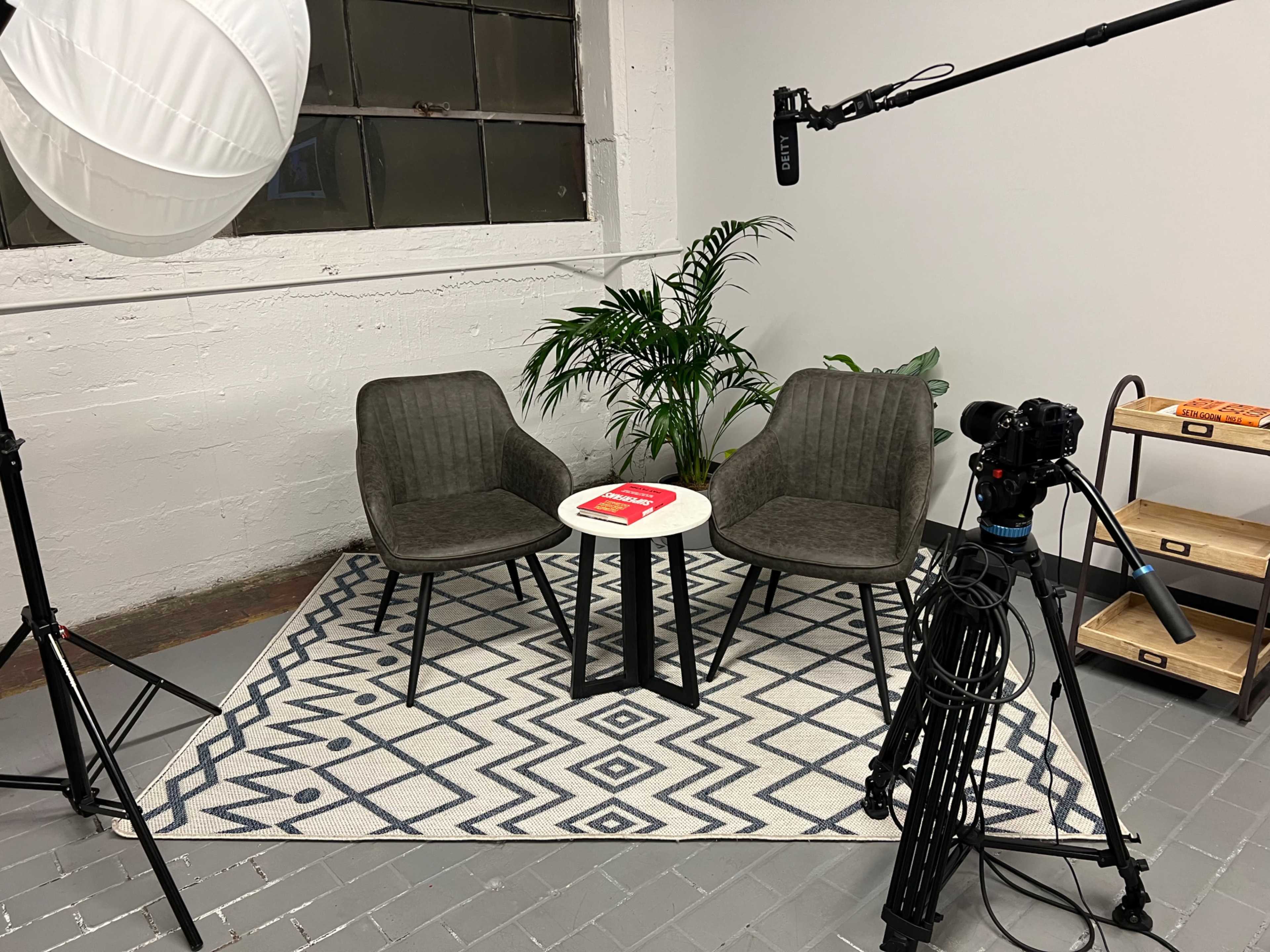 A minimalistic interview setup featuring two chairs facing a small table on a patterned rug, with video equipment and studio lighting nearby.