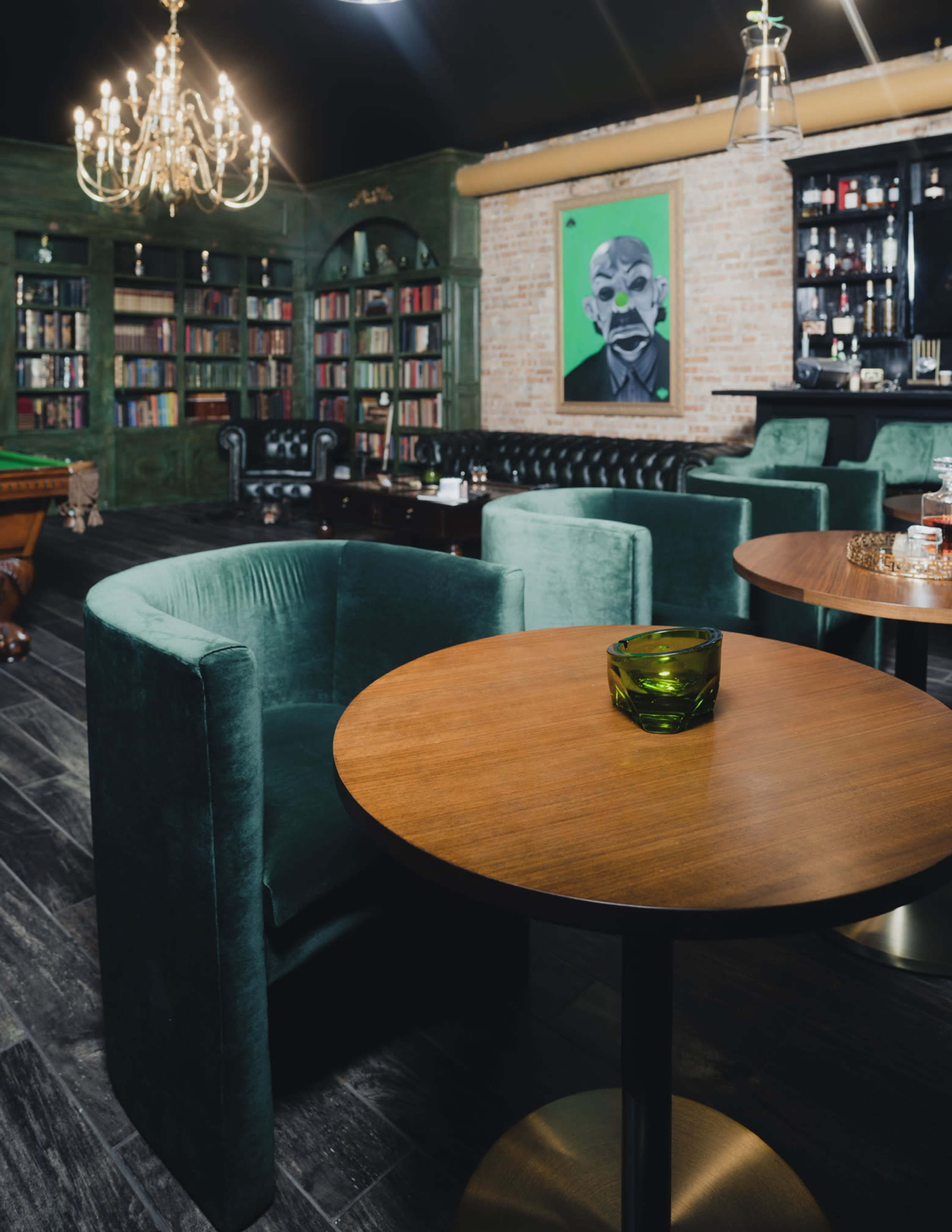 A stylish interior of a bar or lounge with green upholstered chairs, wooden tables, a chandelier, bookshelves, and a vibrant artwork on the wall.