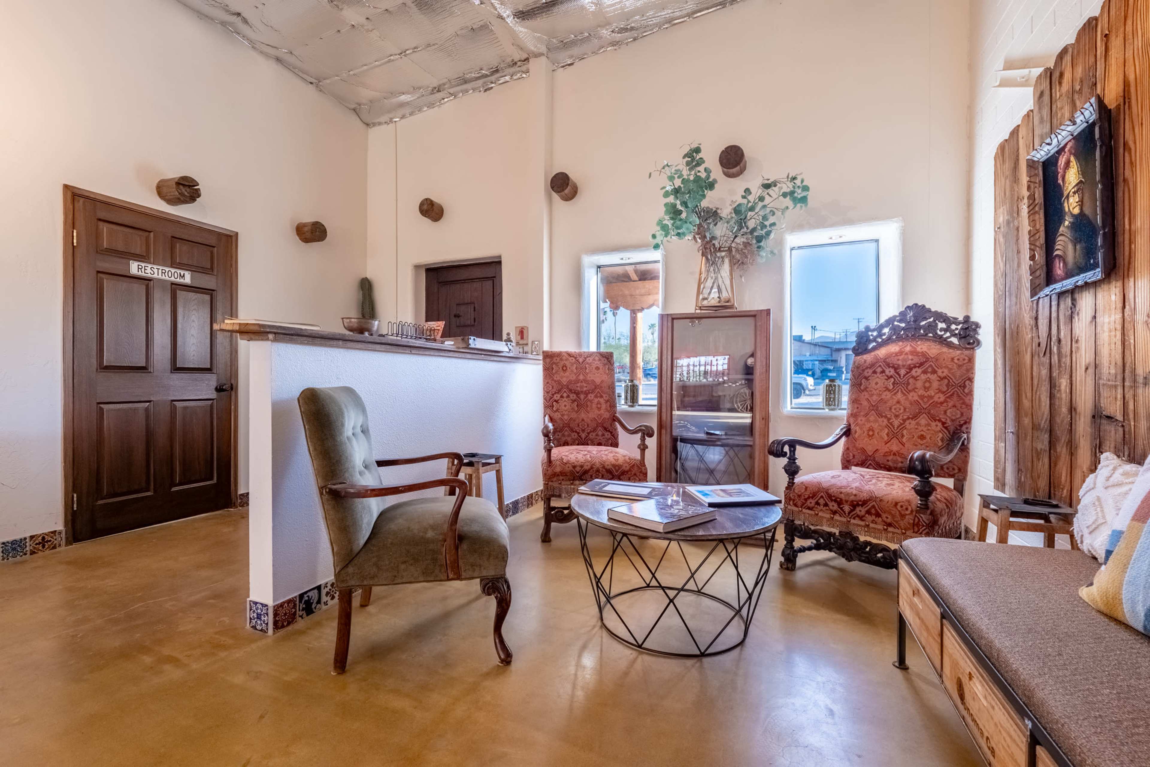 The image shows a cozy waiting area with vintage chairs, a glass coffee table, and a wooden door labeled "RESTROOM."