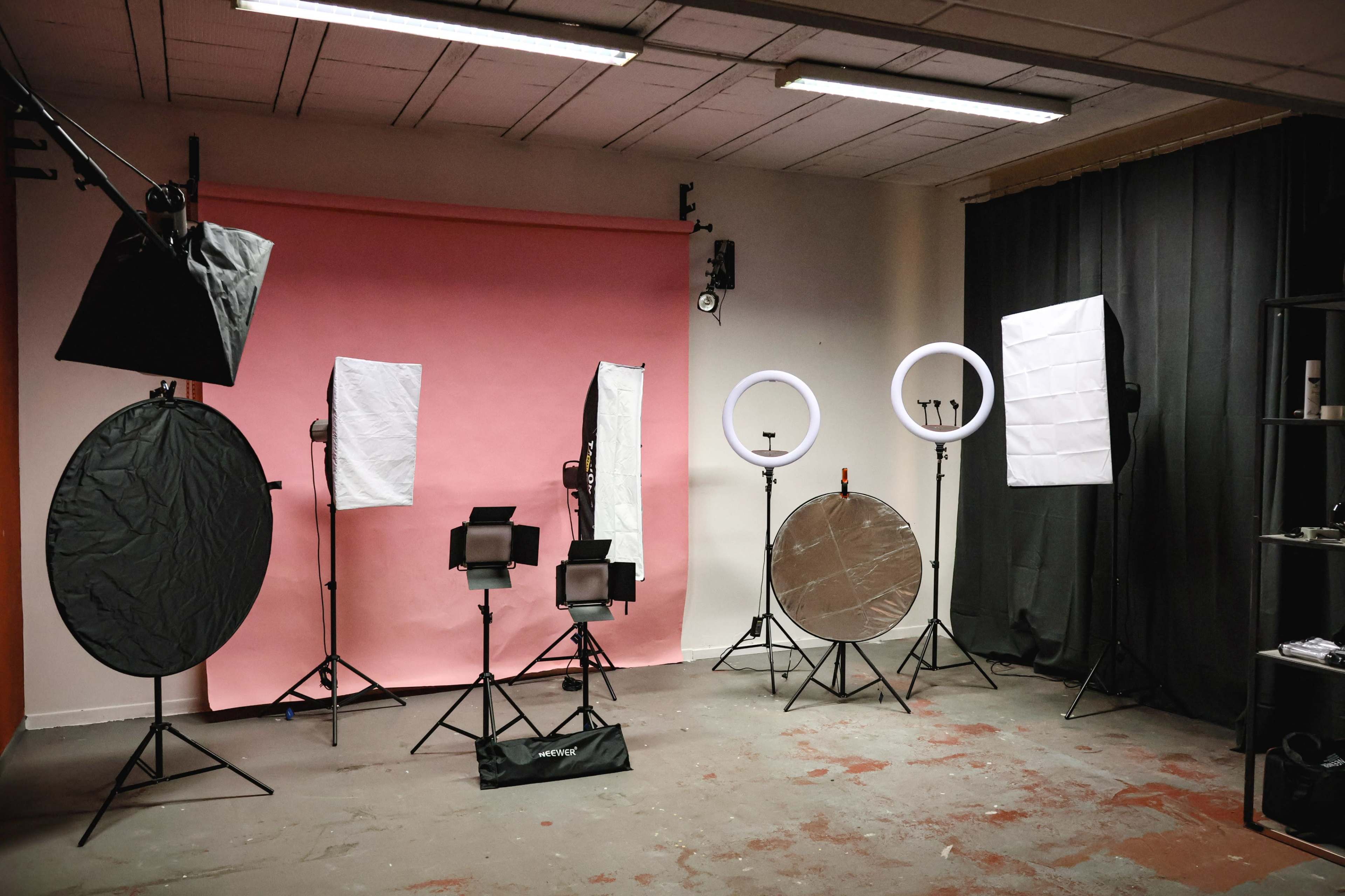 The image shows a photography studio set up with various lighting equipment and backdrops, including a pink backdrop, softboxes, reflectors, and light stands arranged around the room.