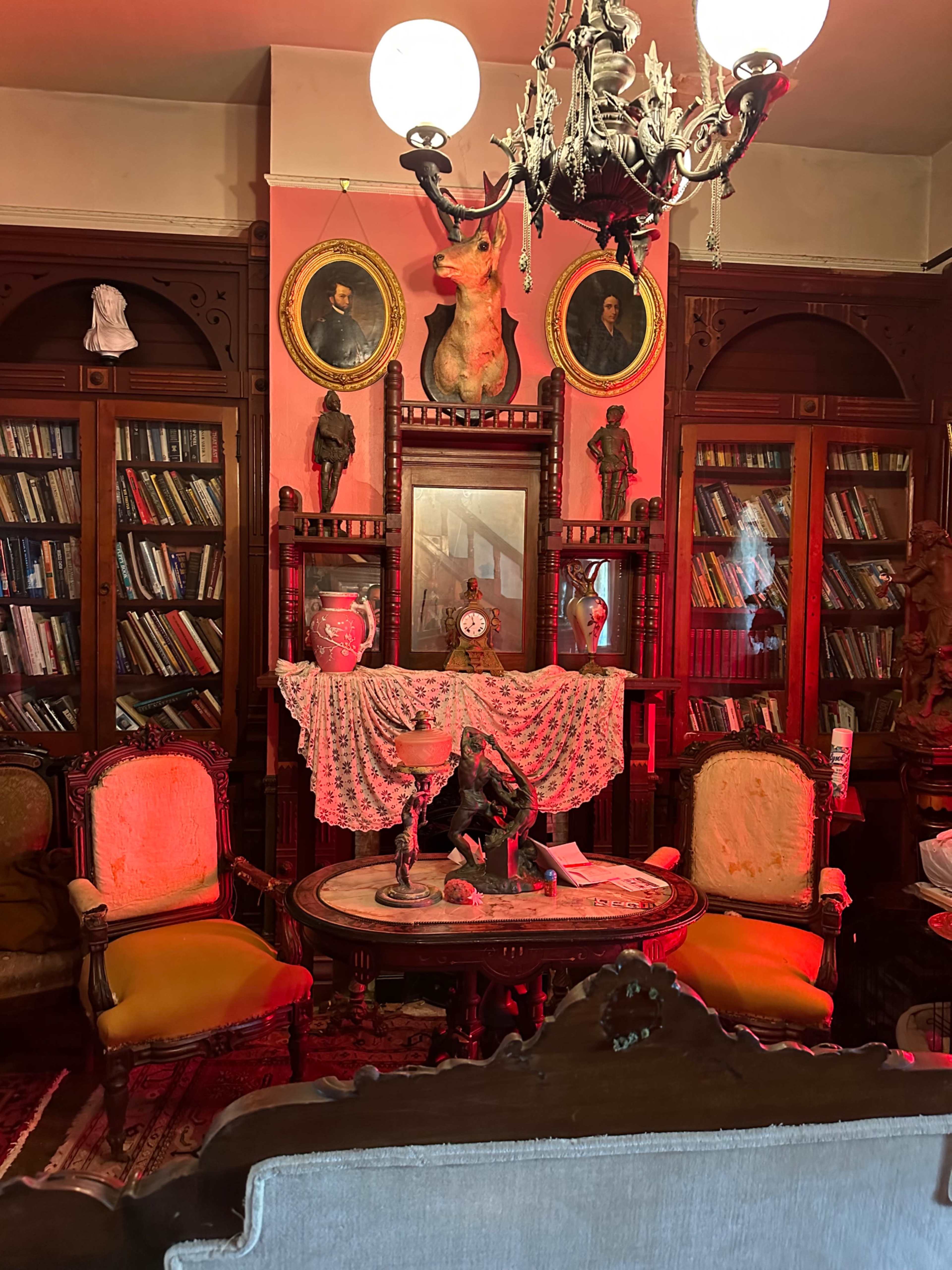 A vintage library with ornate wooden shelves filled with books, a table displaying sculptures, and a chandelier overhead, surrounded by antique chairs.