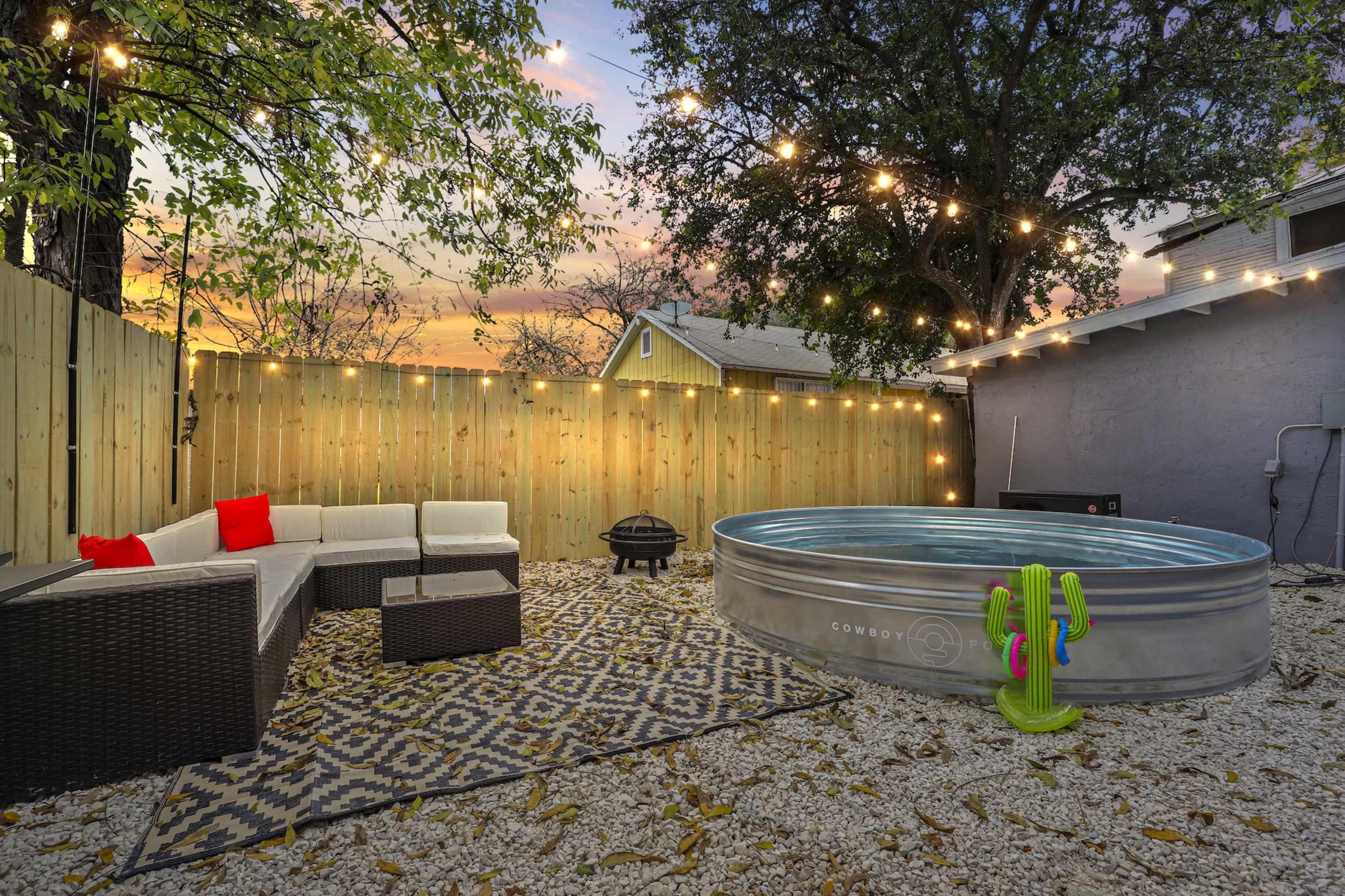 The image shows a cozy outdoor space featuring a circular metal pool, a seating area with a sectional sofa, a decorative rug, and string lights overhead against a sunset sky.