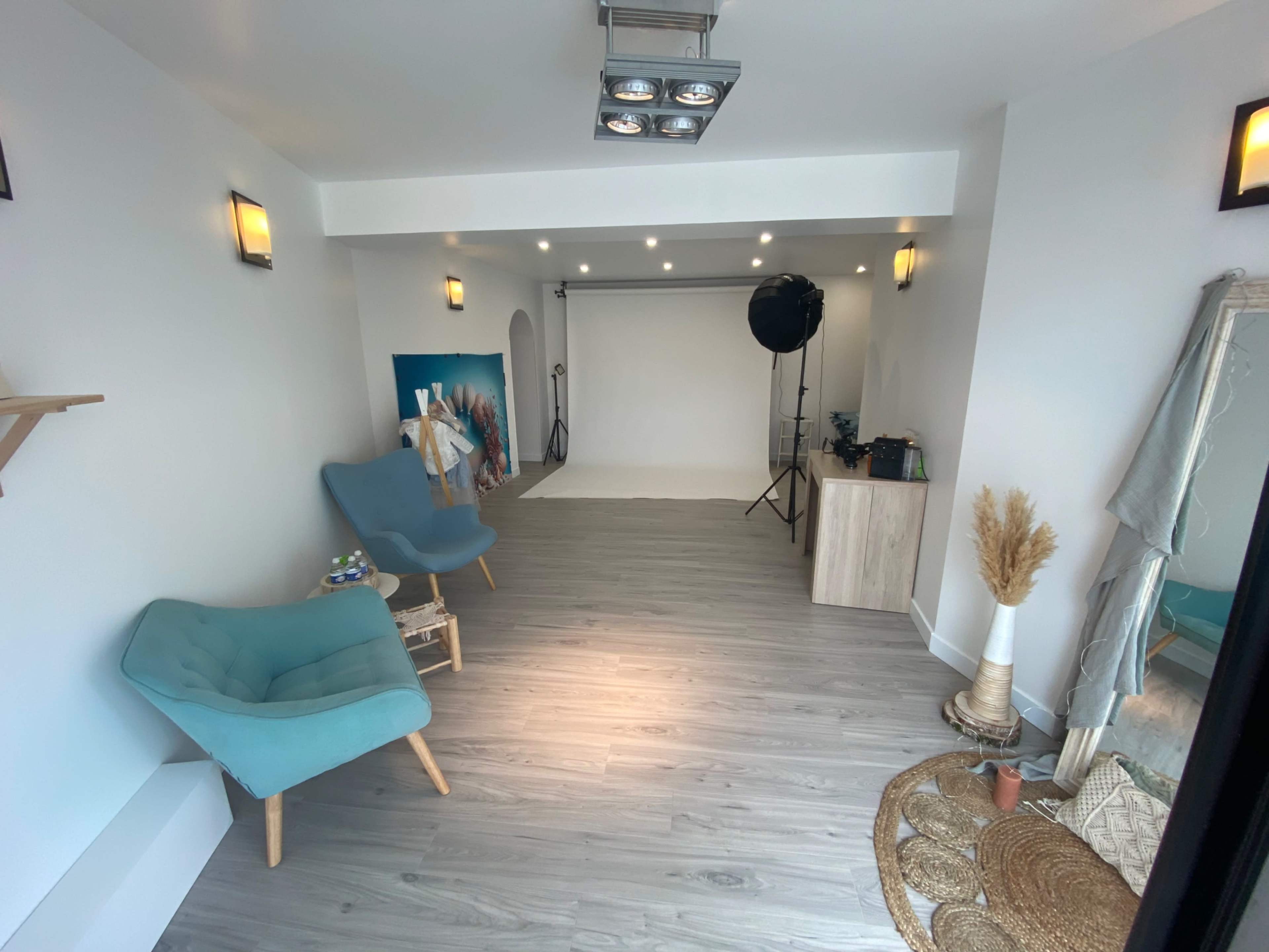 A bright, minimalistic studio space with wooden flooring, two blue chairs, a backdrop setup, and various photography equipment arranged neatly.
