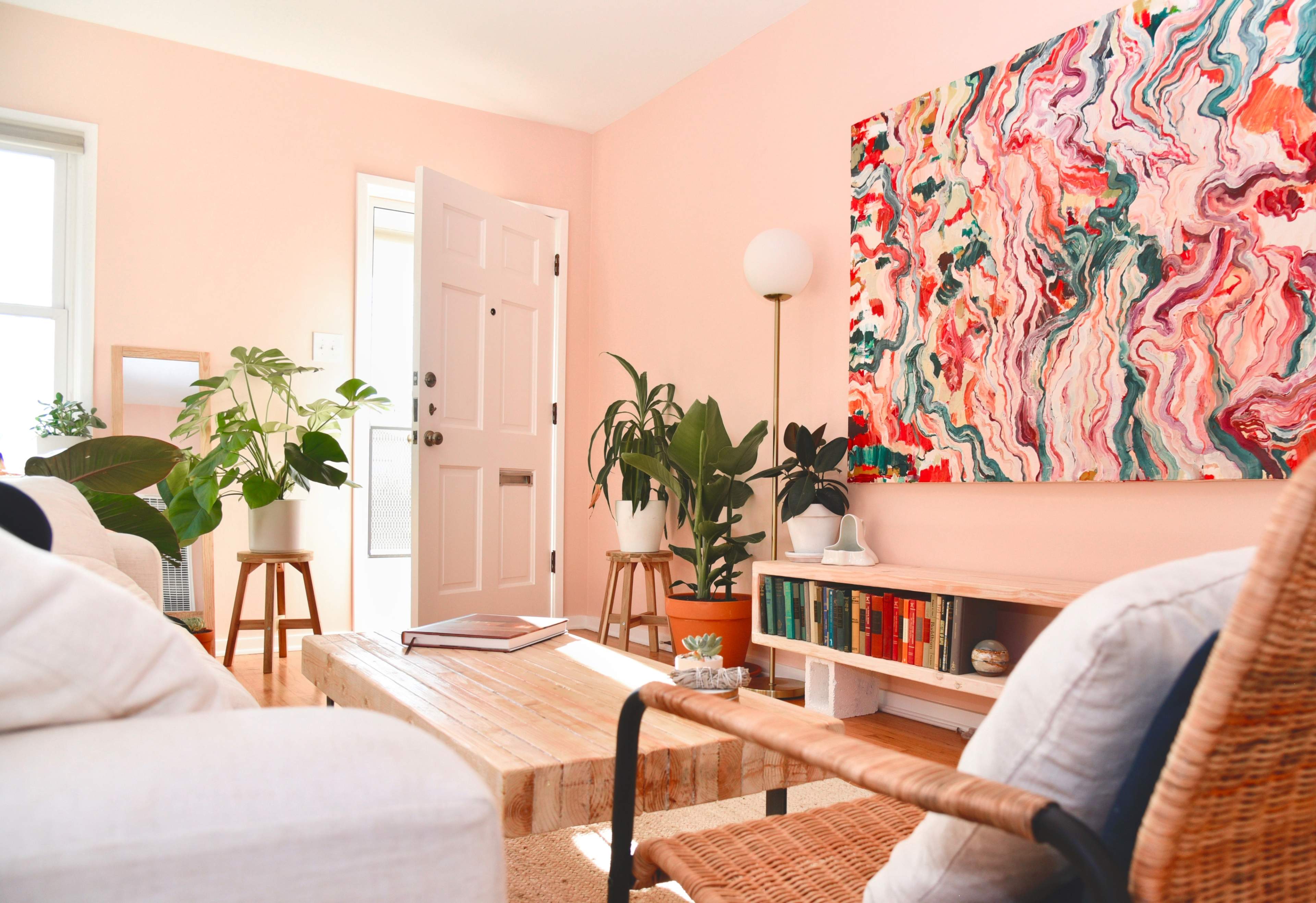 A brightly lit living room featuring a pink wall, a large colorful abstract painting, potted plants, a wooden coffee table, and wicker furniture.