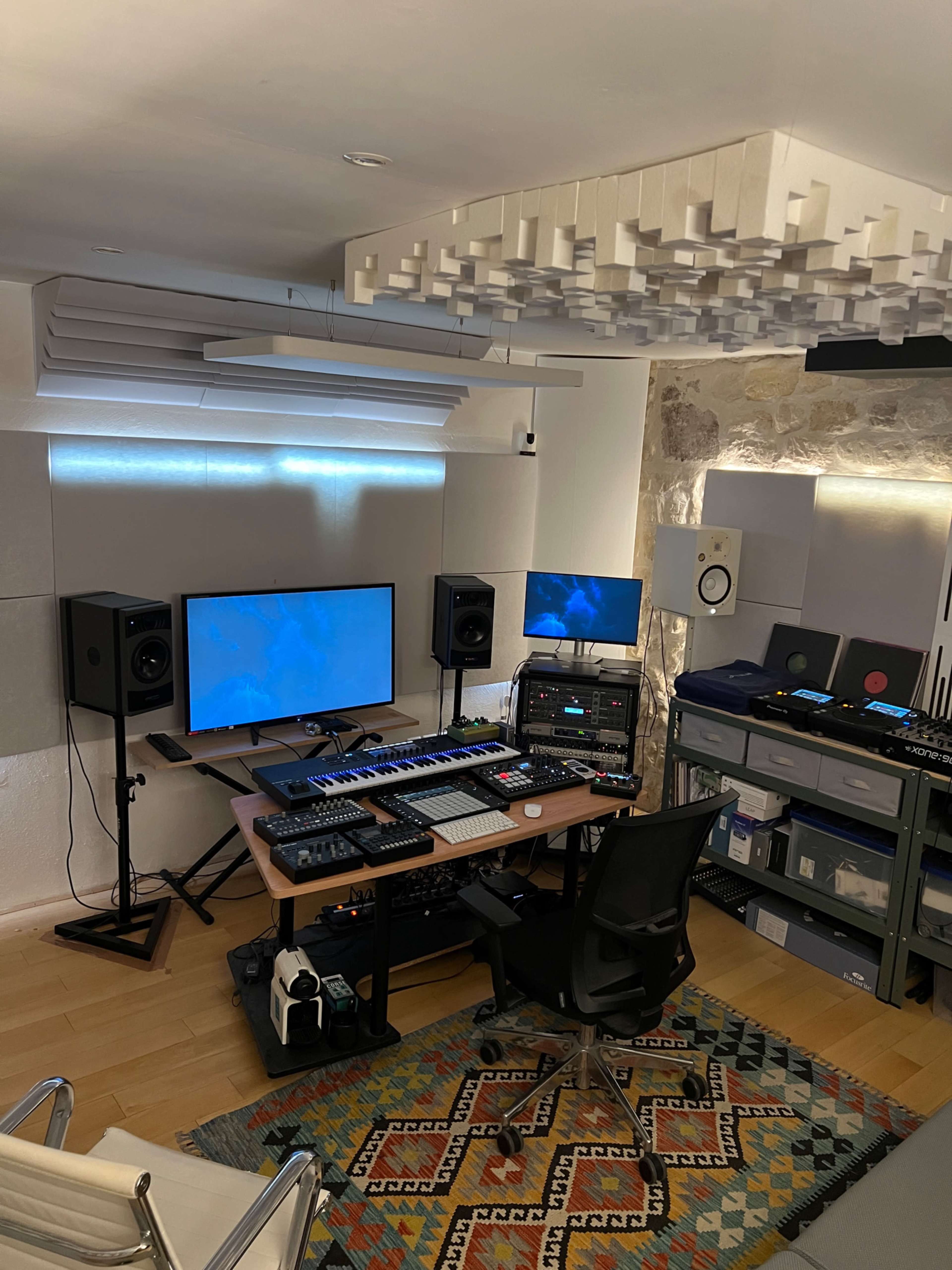 The image shows a music production studio featuring two computer monitors, audio equipment, a keyboard, and shelves with various electronic devices, all set against a textured wall with soundproofing panels.
