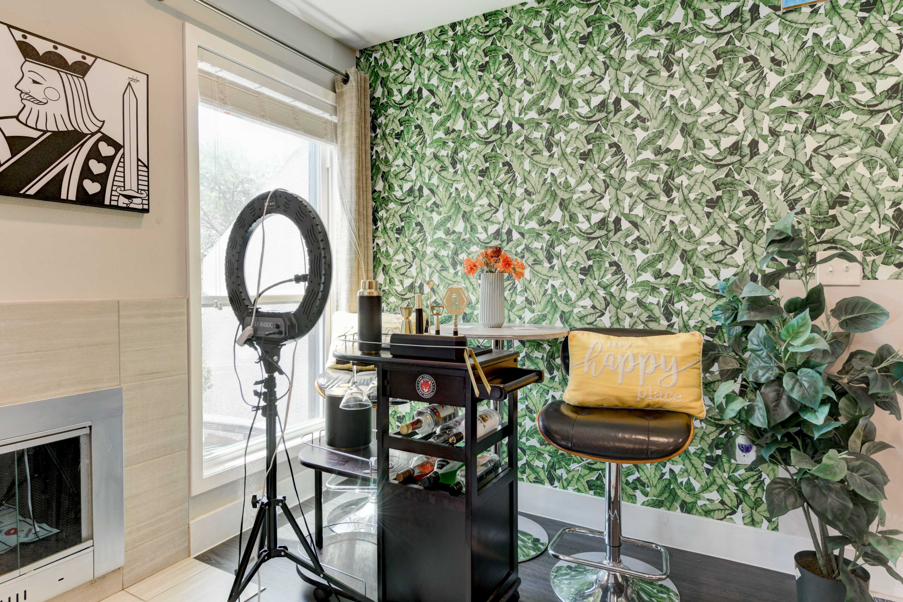 A modern corner of a room with a bar cart, a ring light, a chair, and decorative plants against a leafy wallpaper backdrop.