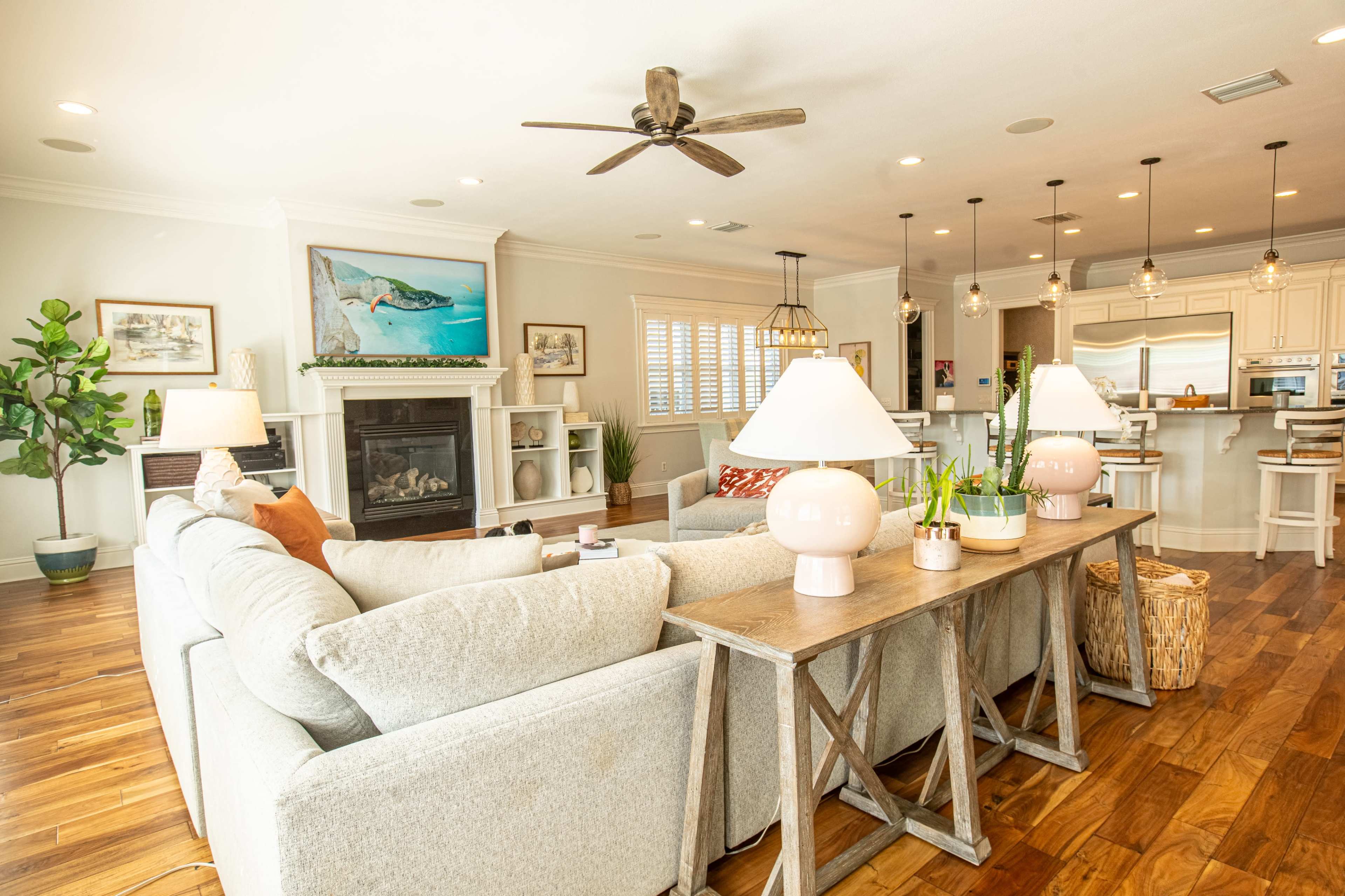 The image shows a spacious living room featuring a light-colored sectional sofa, a wooden console table with lamps, a fireplace, and an open kitchen area in the background.