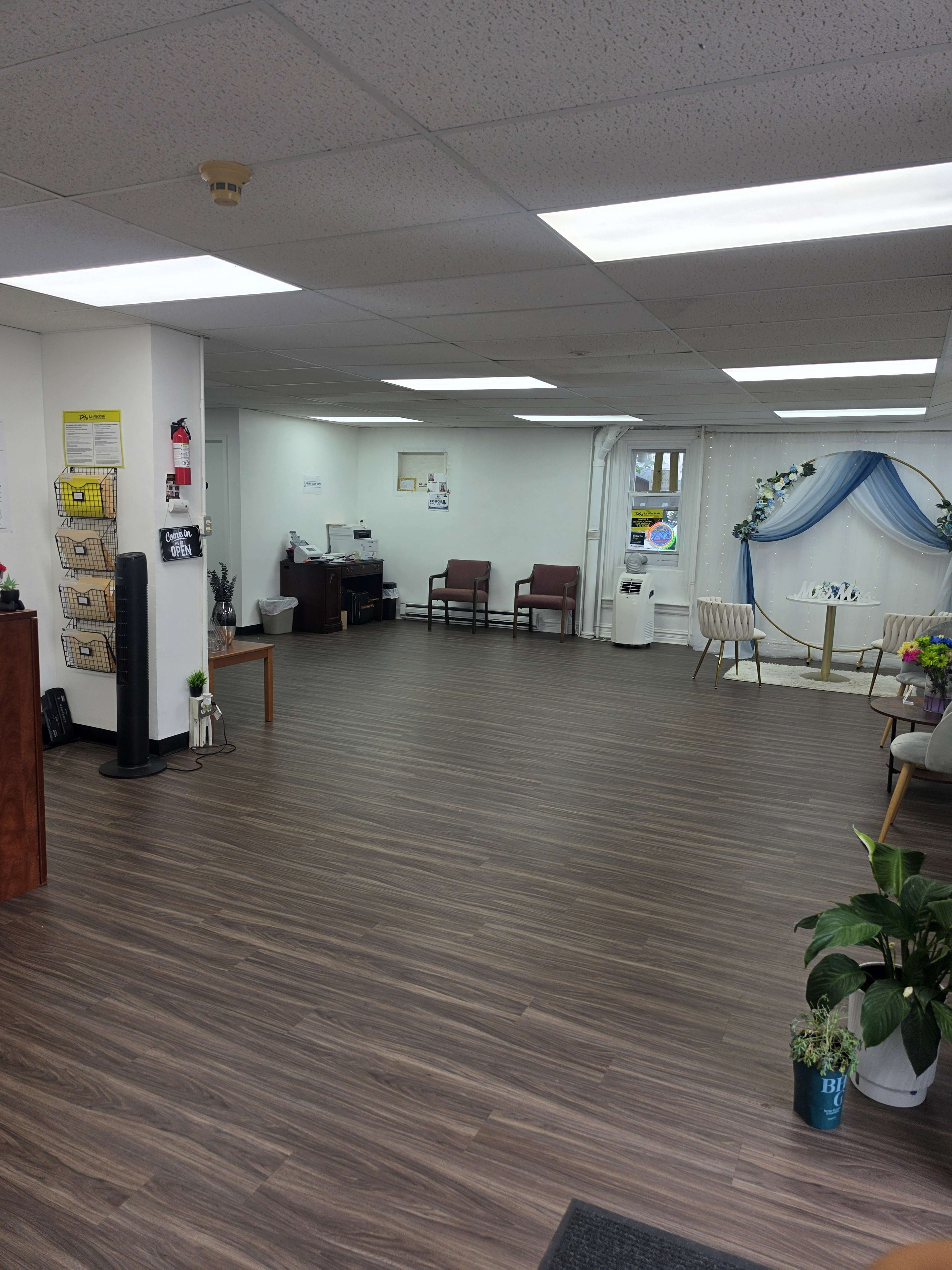 The image shows a spacious waiting area with wooden flooring, several chairs arranged in a circle, and a small decorative setup featuring a blue draped backdrop.