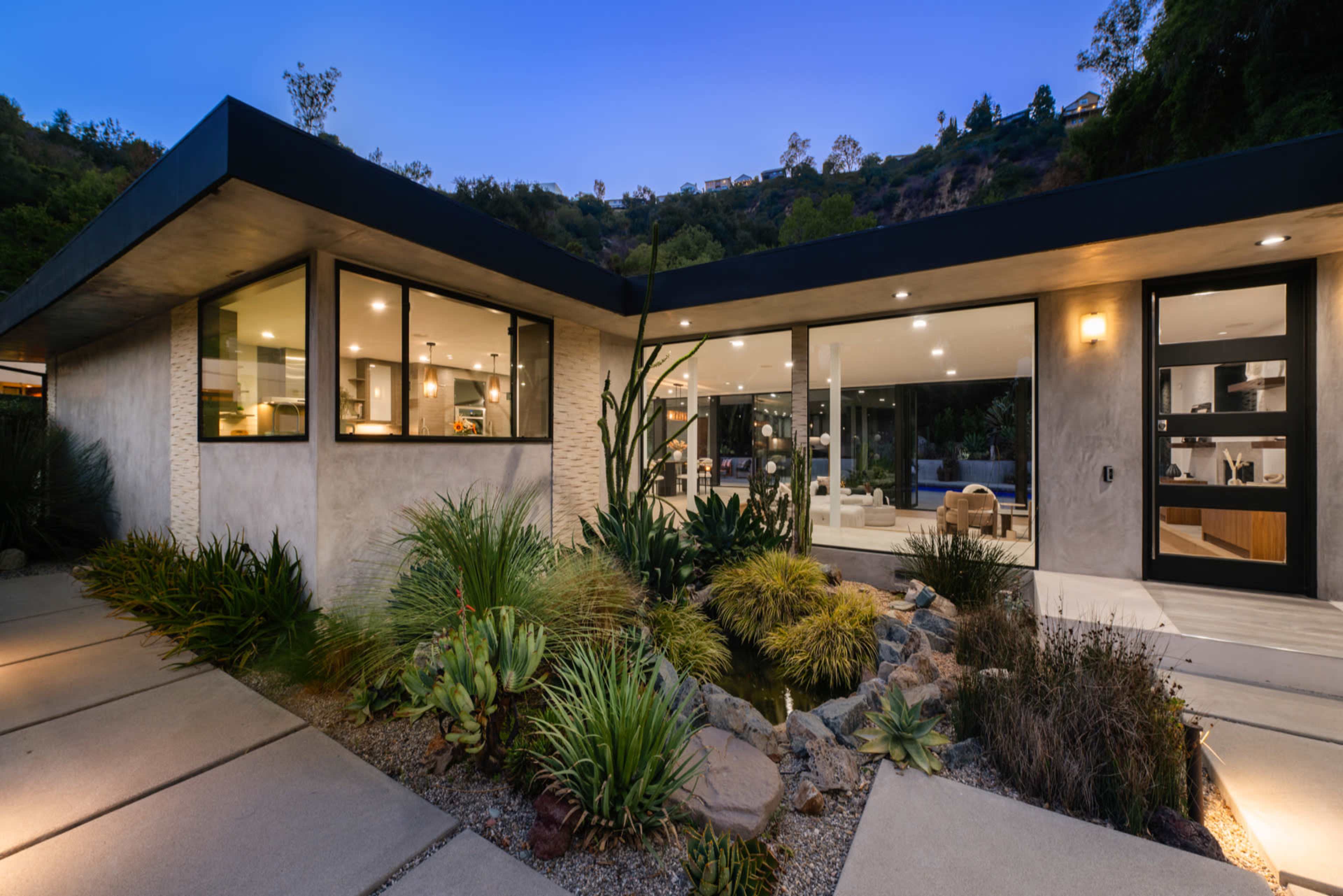 A modern house features a landscaped entrance with various plants and a water feature, illuminated under evening lighting.