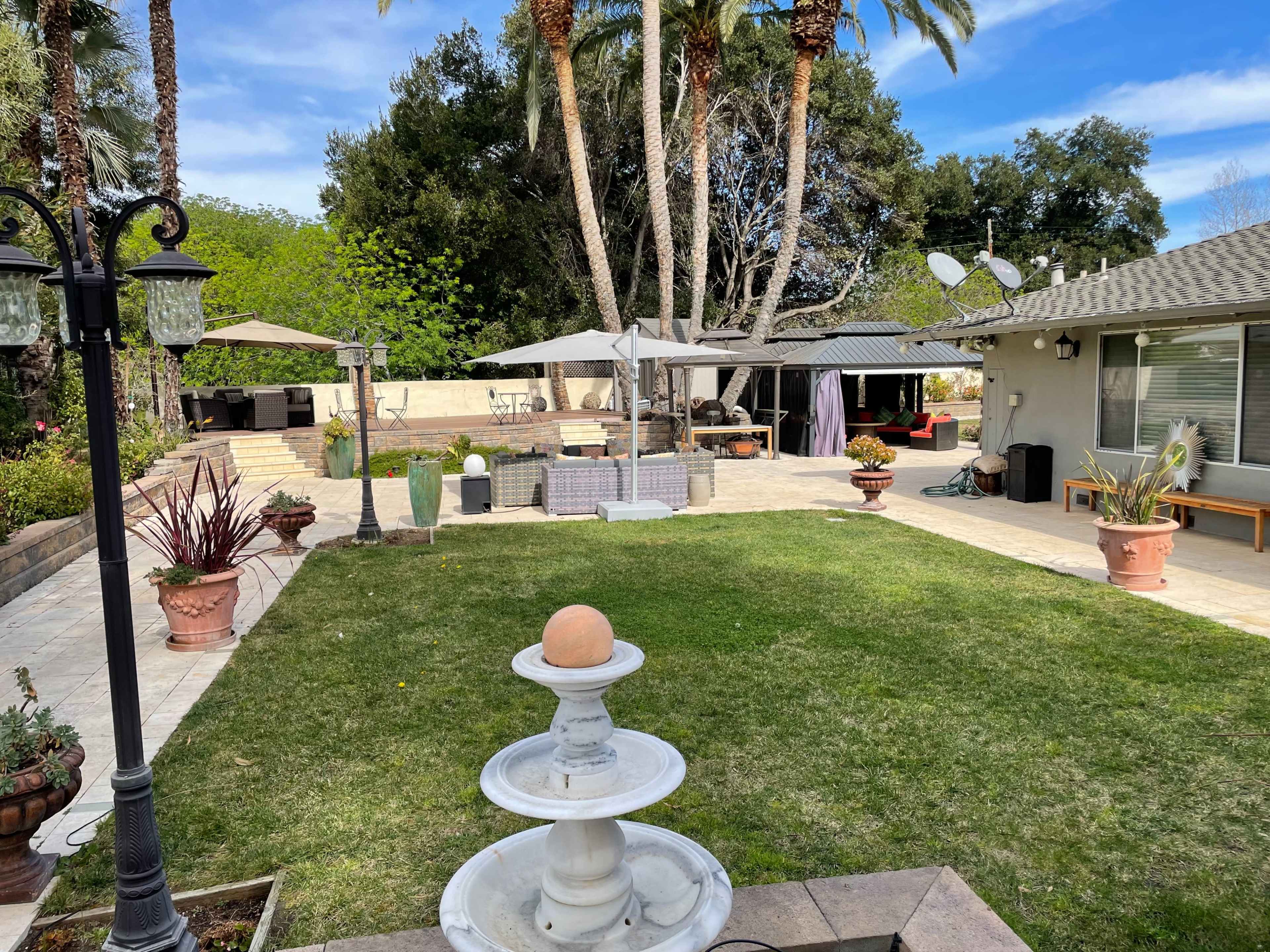 The image shows a landscaped backyard featuring a fountain, several potted plants, outdoor seating areas, and palm trees against a clear blue sky.
