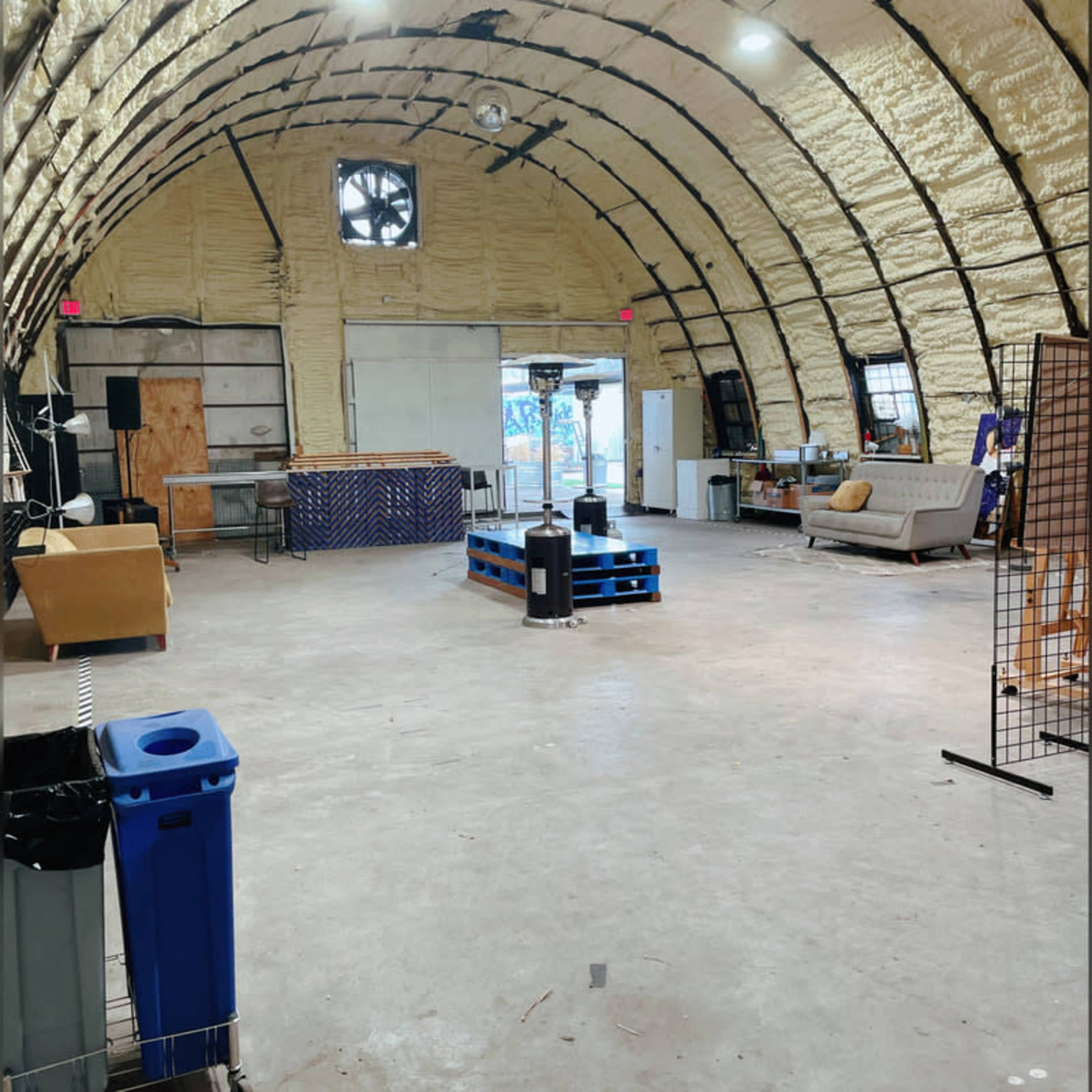 A spacious, empty indoor area with a curved ceiling, featuring seating, a small table on pallets, and various items along the walls.
