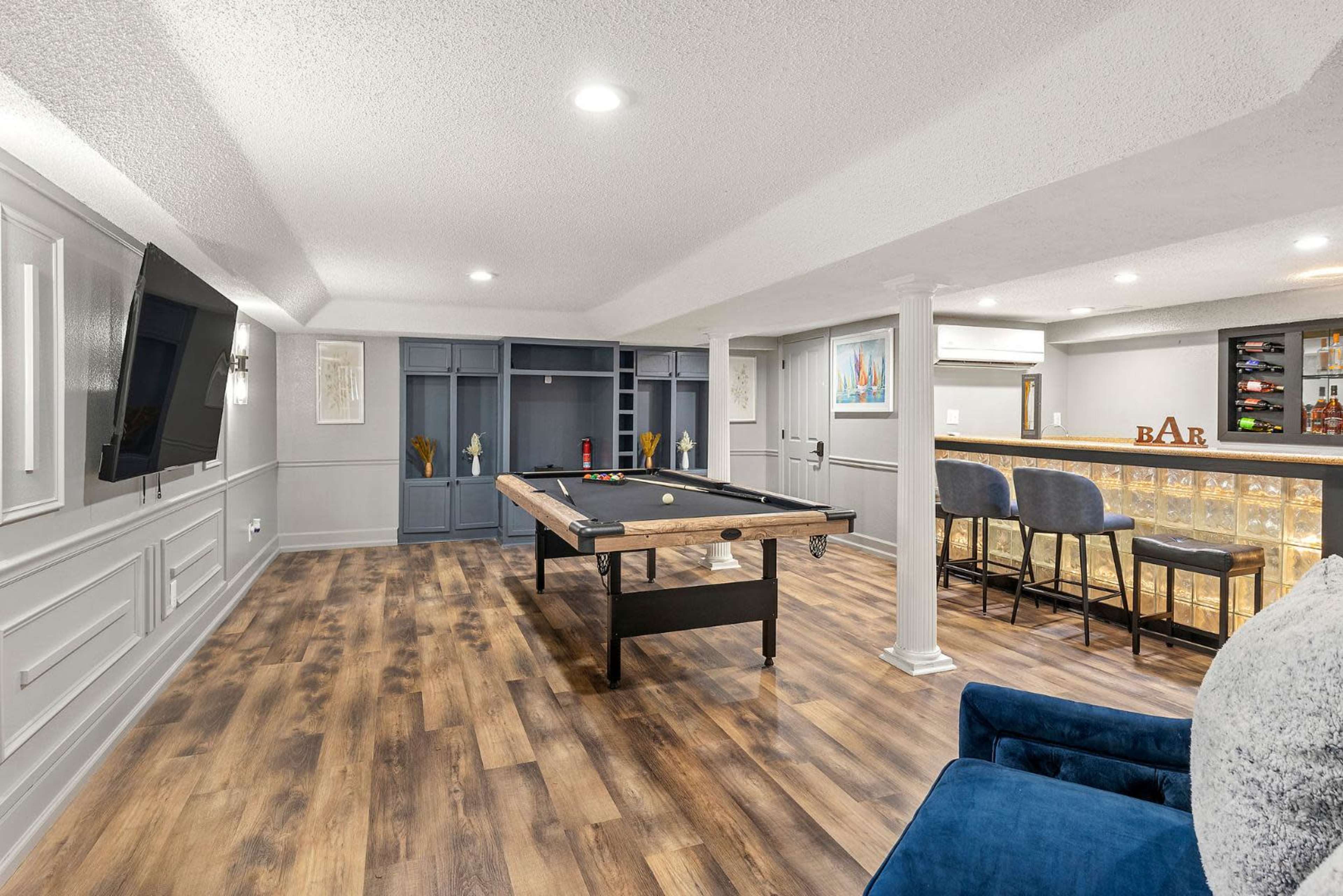 The image shows a modern basement game room featuring a billiards table, a bar area with seating, and built-in shelving.