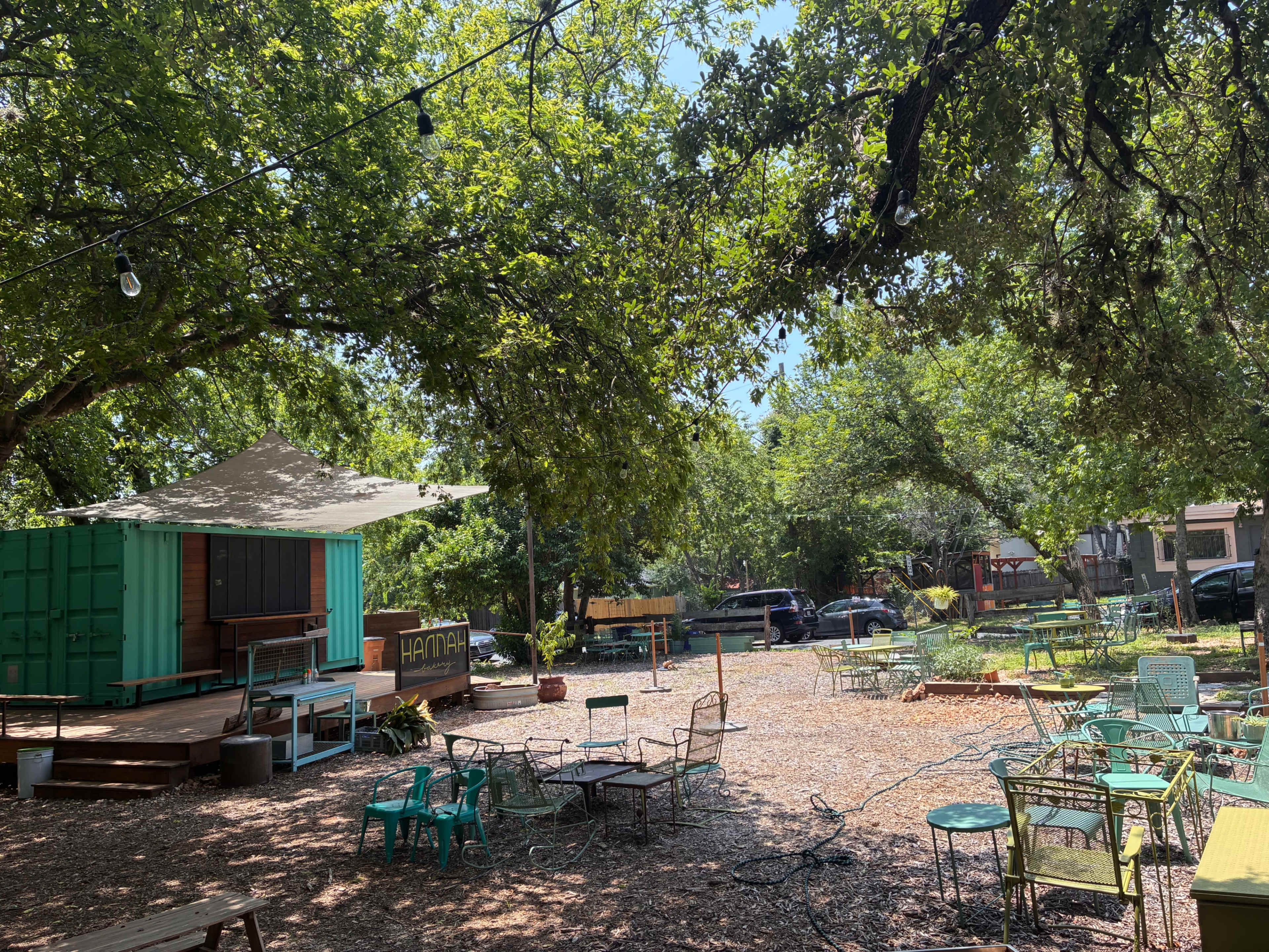 The image shows an outdoor seating area with green chairs and tables under trees, near a colorful building or stage.