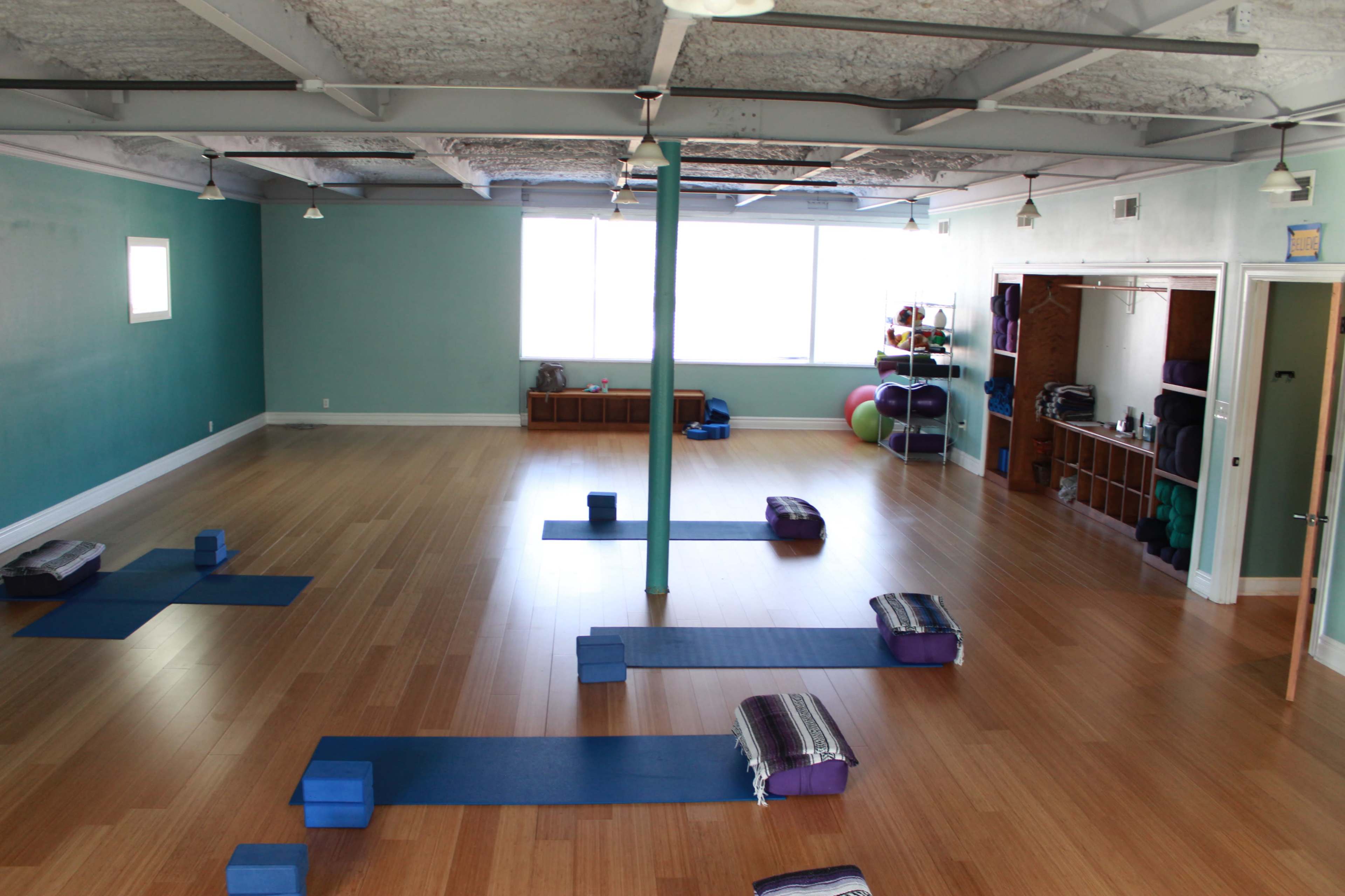 The image shows a spacious yoga studio with wooden floors, blue mats arranged for practice, and a large window letting in natural light.