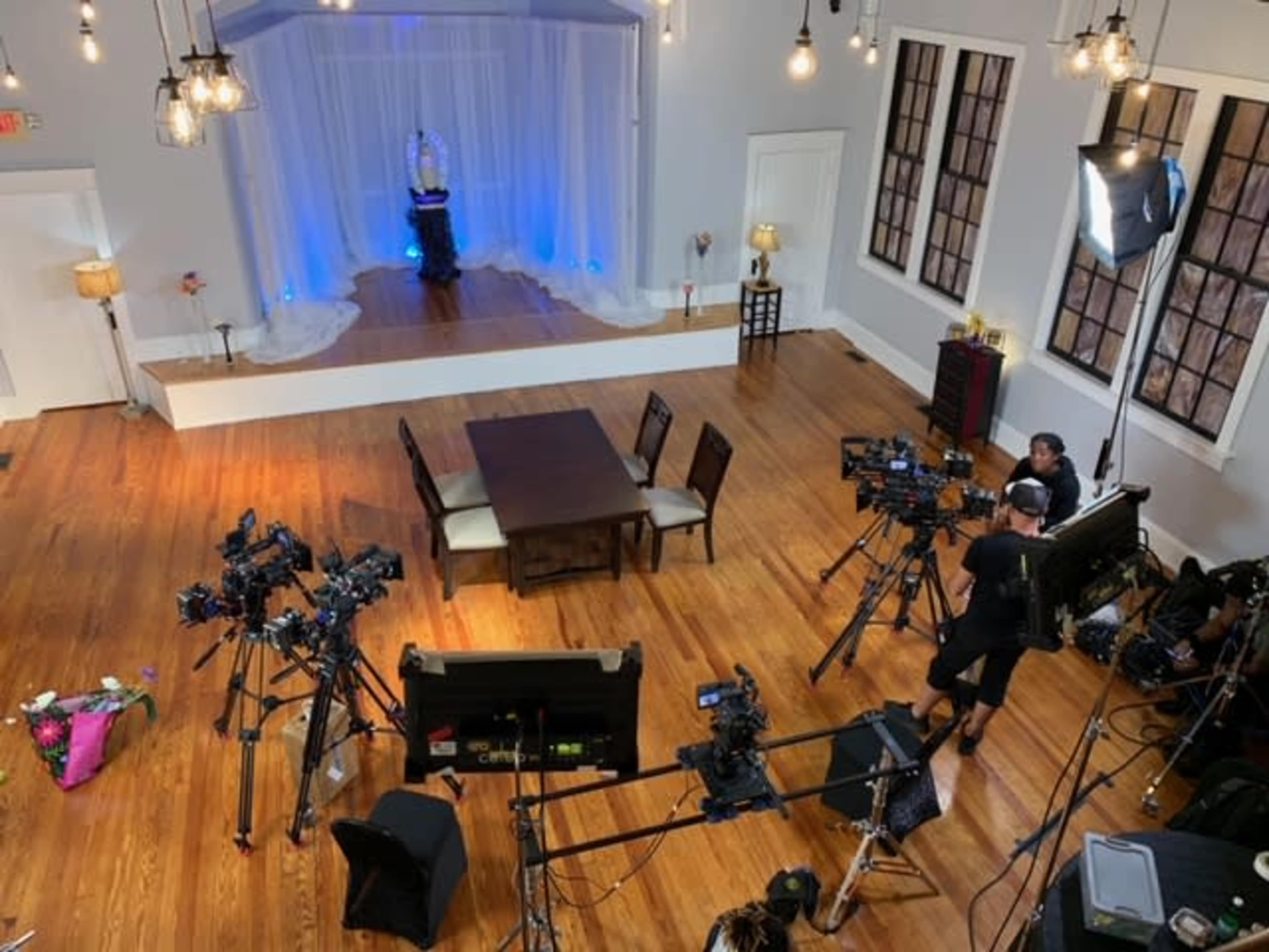 A spacious room set up for a video production, featuring multiple cameras pointed towards a small stage with a decorative backdrop.