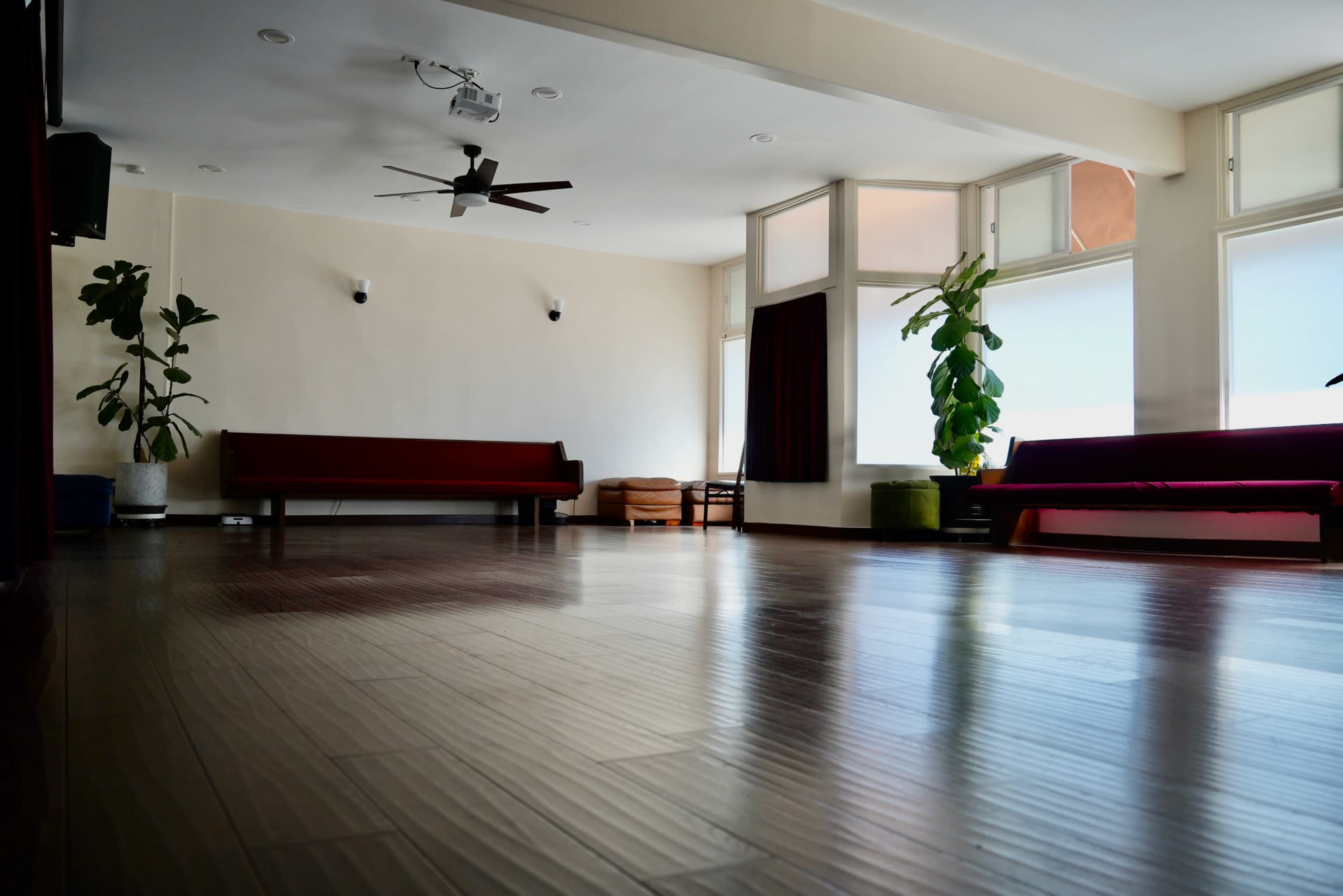 The image shows a spacious room with wooden flooring, featuring benches along the walls and large windows allowing natural light.