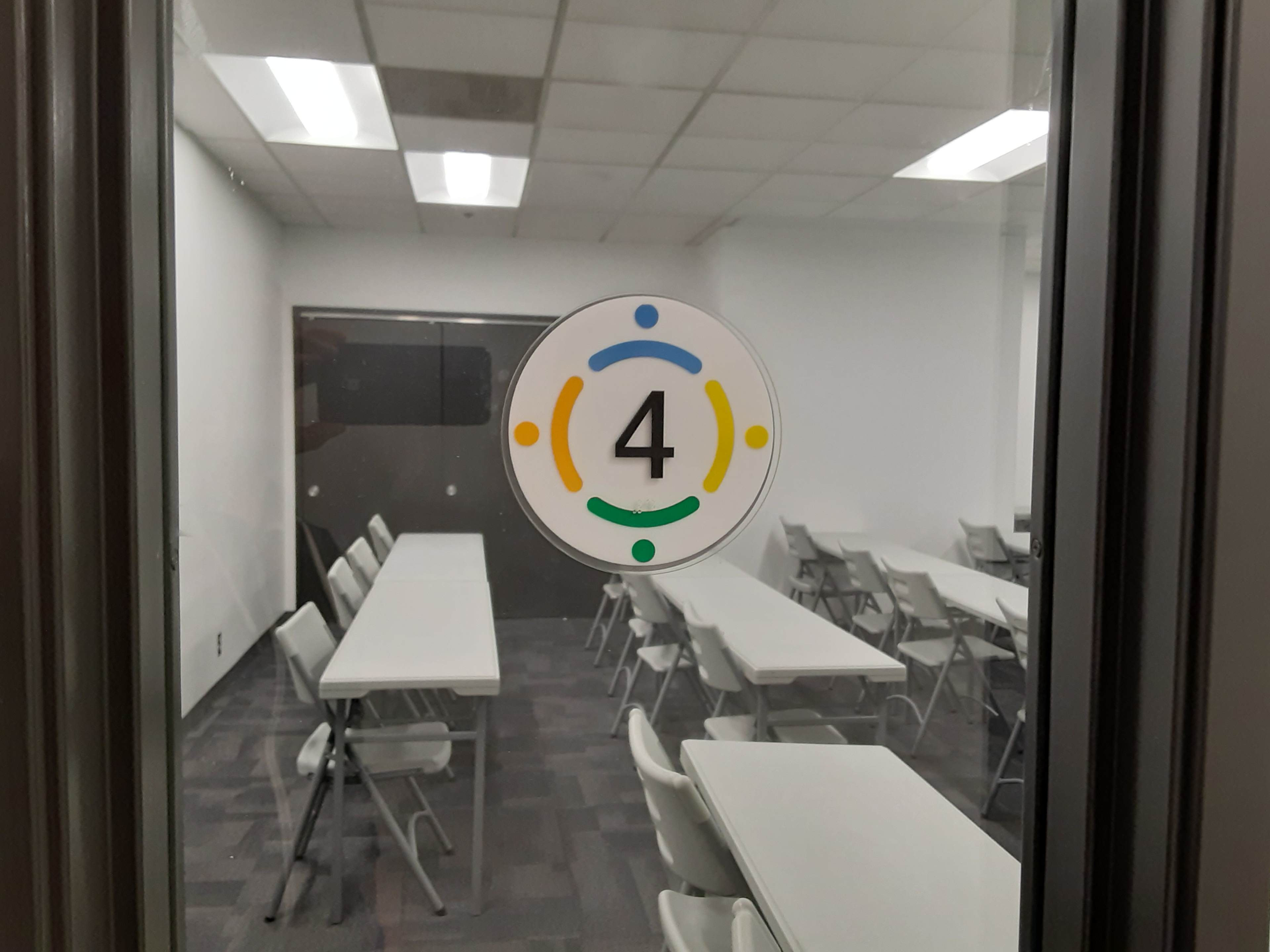 A circular sign displaying the number "4" is affixed to a glass door leading to a room with multiple white tables and chairs arranged for a meeting.