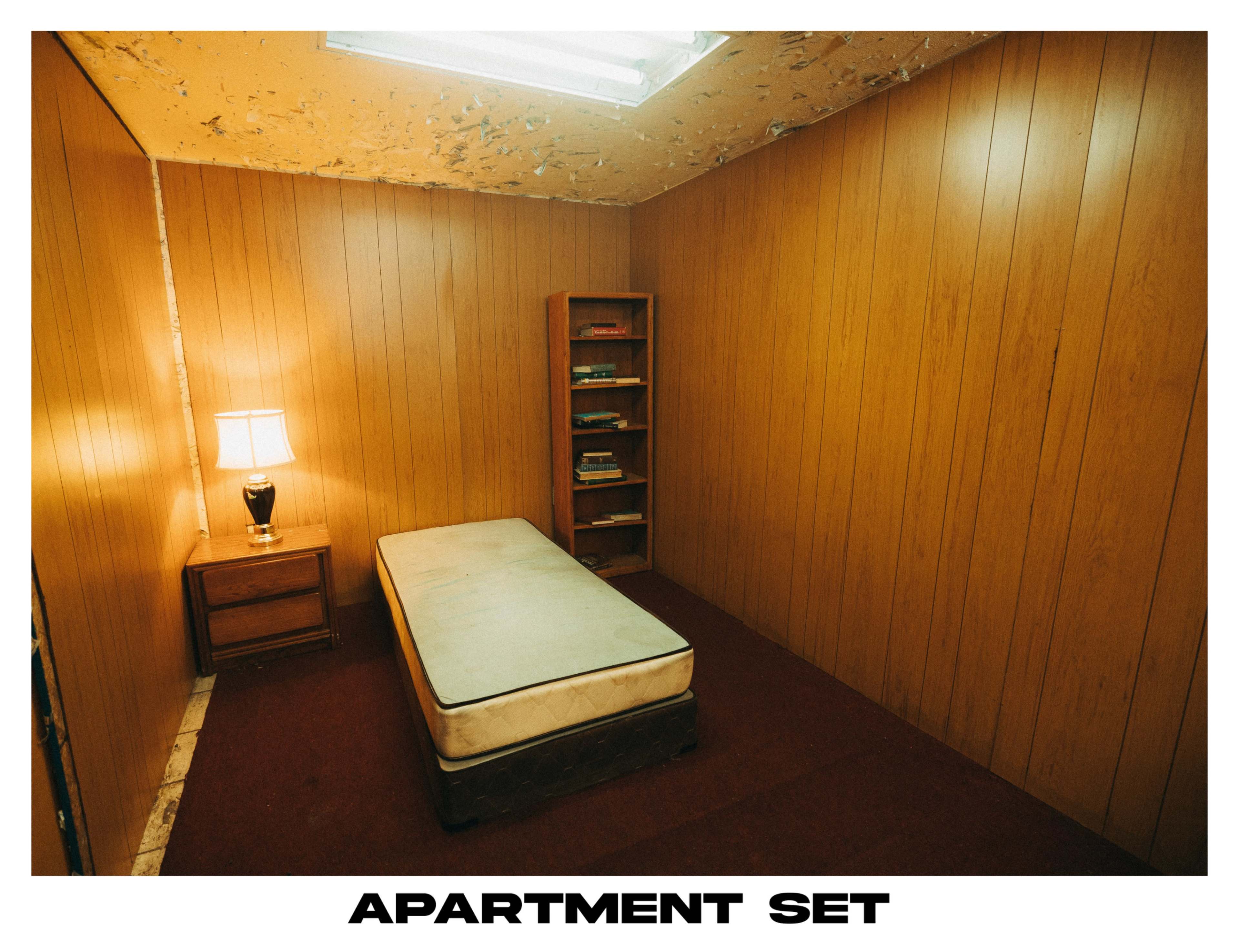 The image shows a sparsely furnished room with wood-paneled walls, a single mattress on the floor, a bedside lamp, and a bookshelf against one wall.