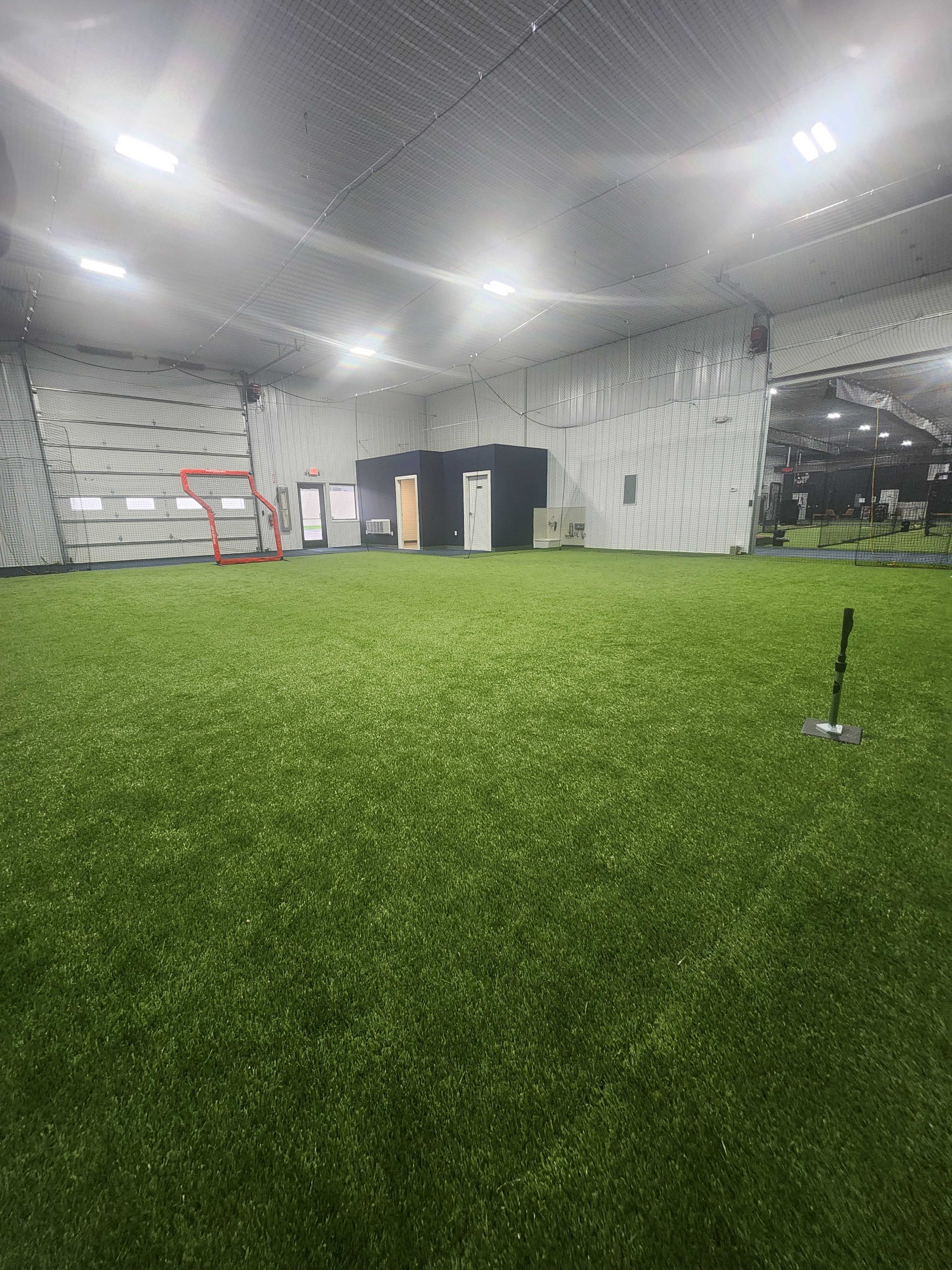 The image shows an indoor facility with artificial turf, featuring a soccer goal at one end and a black storage room along the wall.