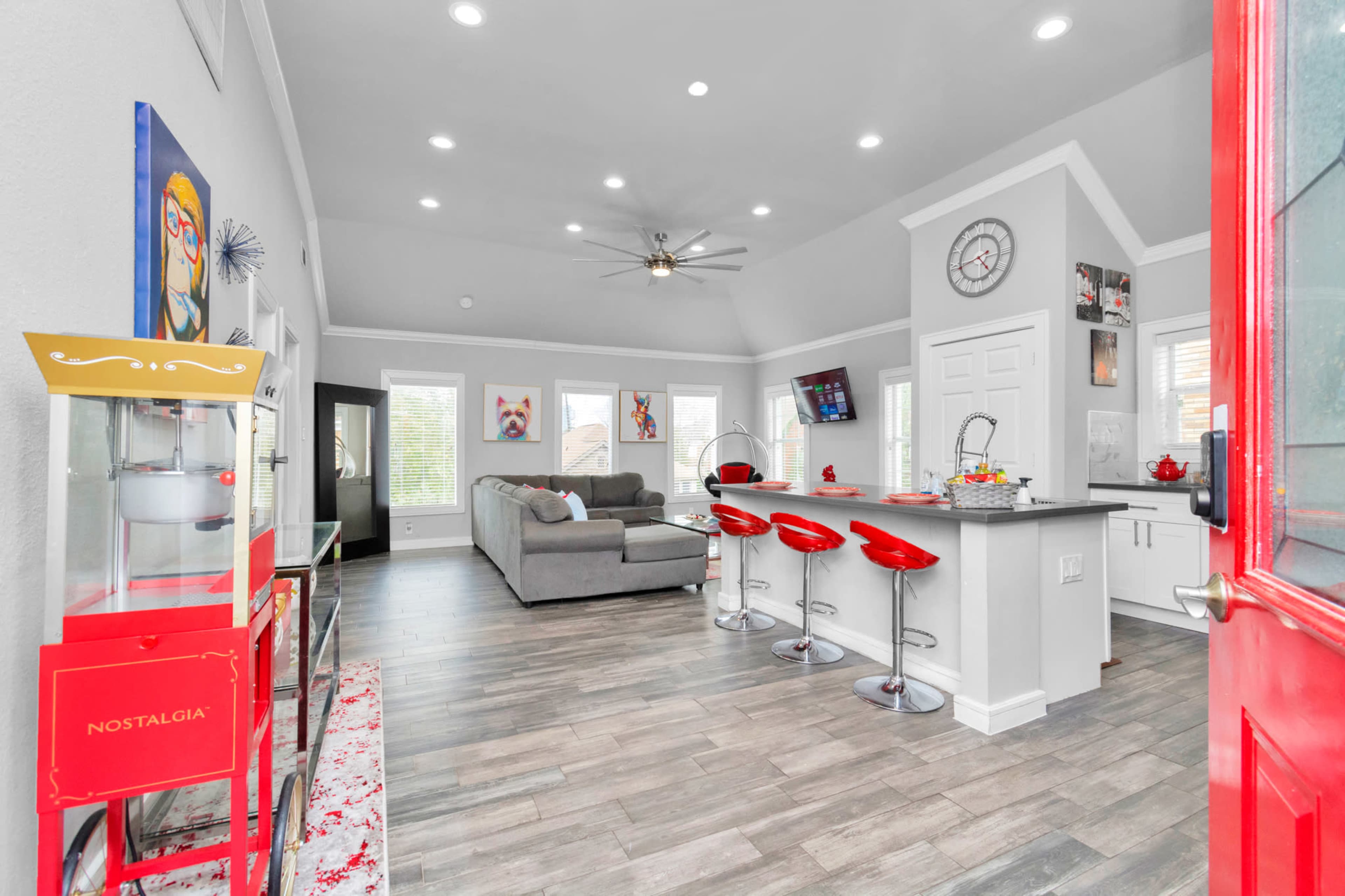 A modern living room and kitchen area with a gray sofa, red bar stools, and large windows illuminating the space.