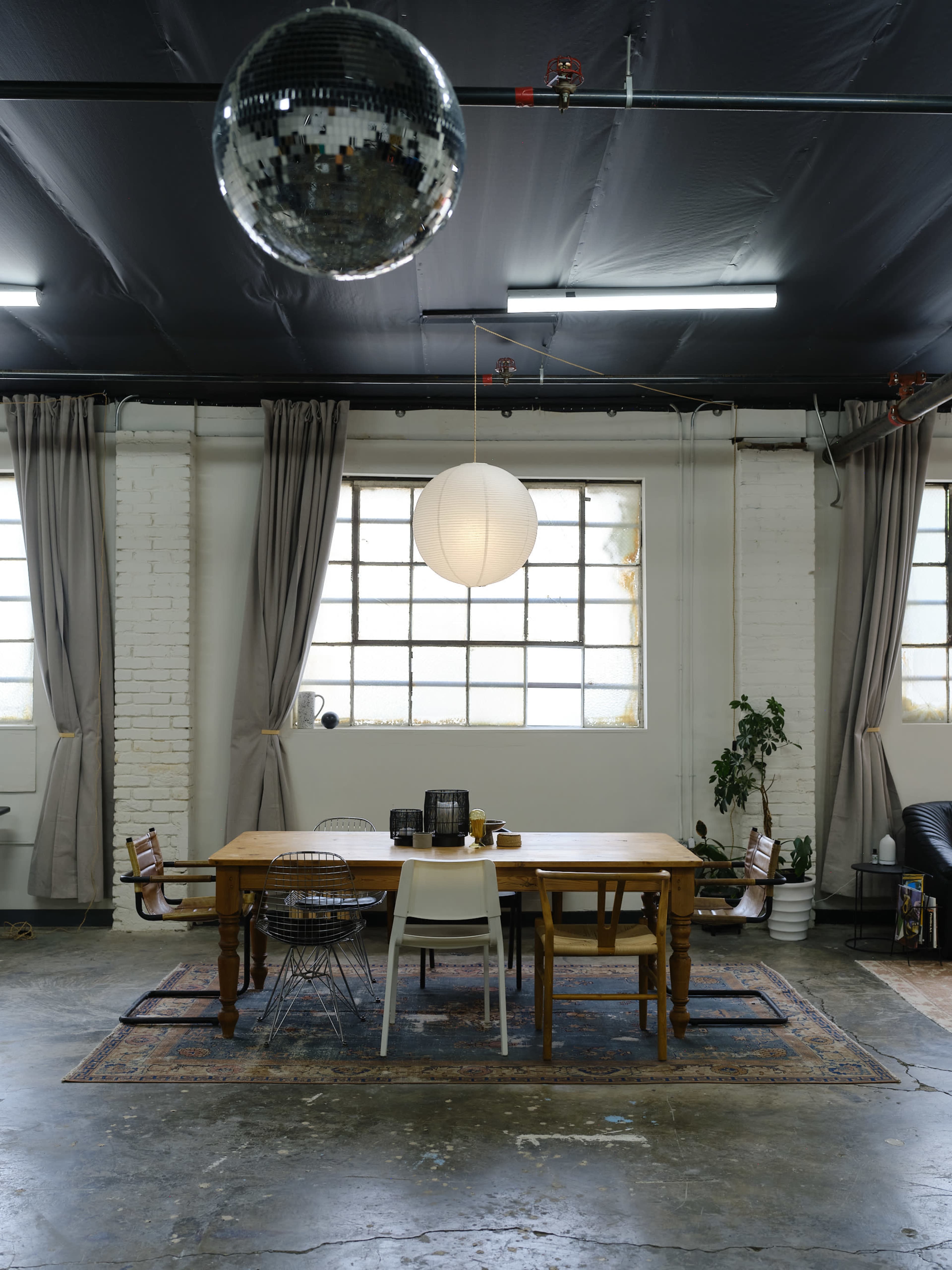 A spacious, industrial-style room featuring a large wooden dining table surrounded by metal and plastic chairs, with a disco ball hanging from the ceiling.