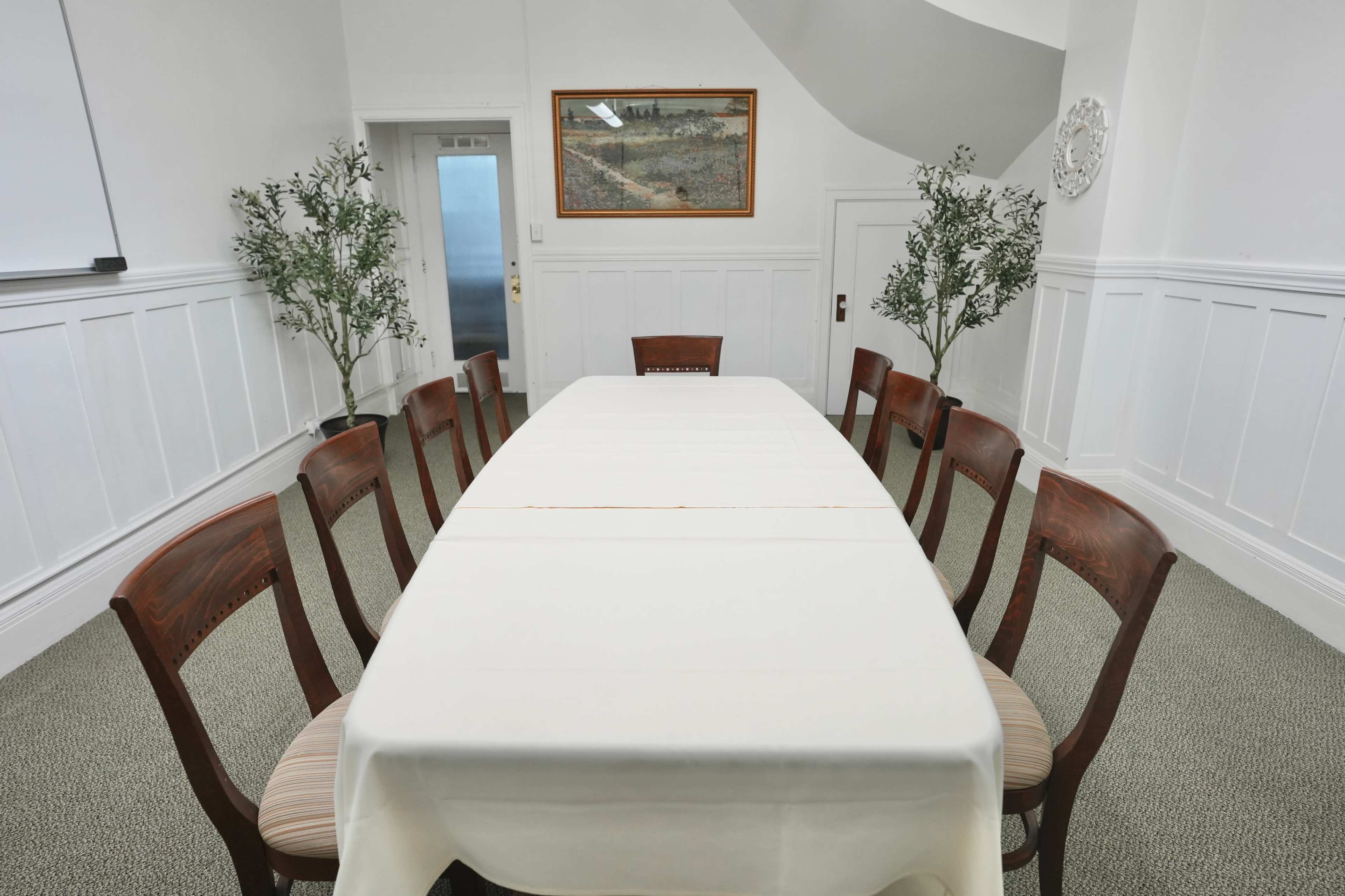 A long table covered with a white tablecloth is set in a bright room with wooden chairs arranged around it, flanked by potted plants.