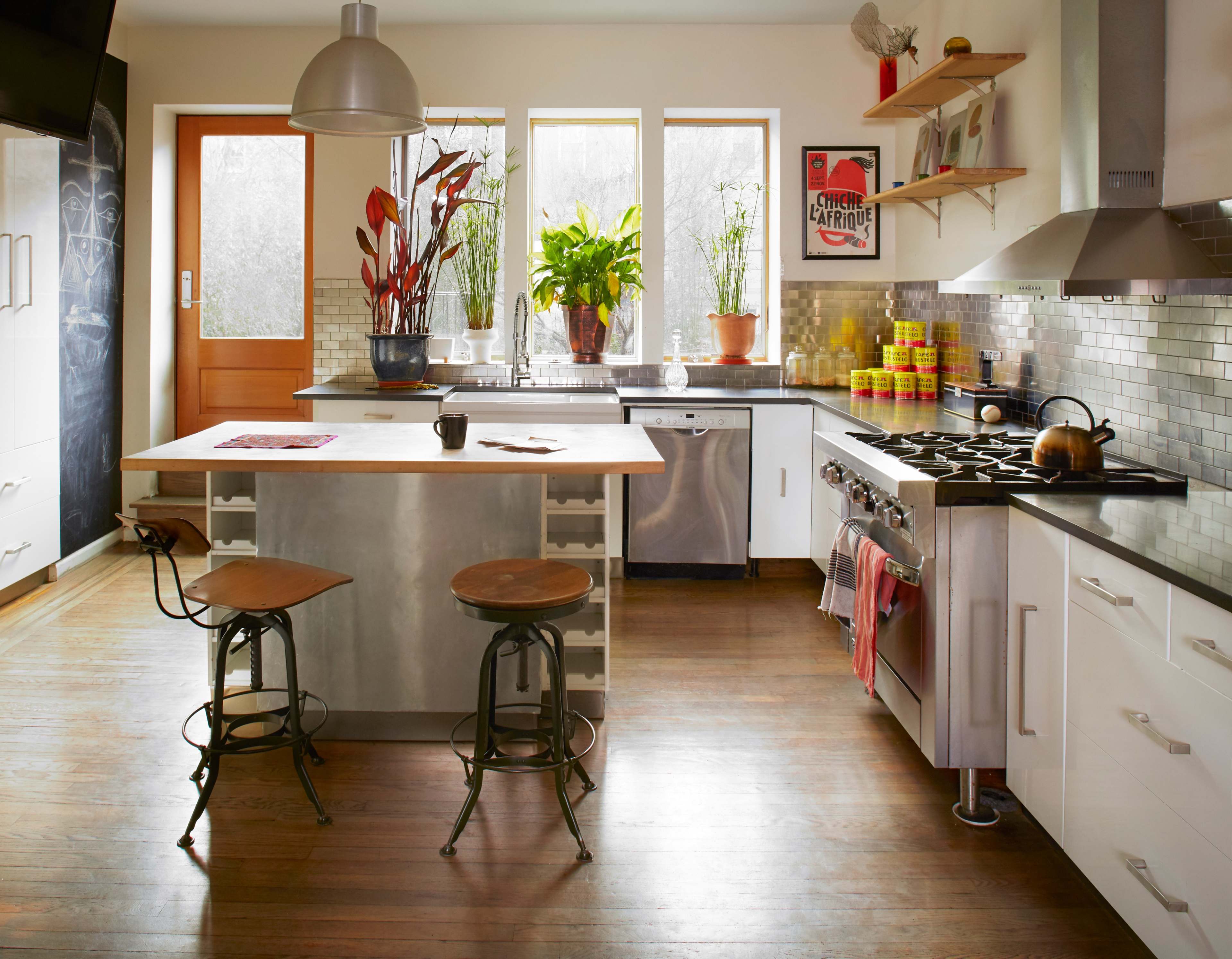 A modern kitchen features a central island with metal seating, stainless steel appliances, and large windows allowing natural light to illuminate the room.