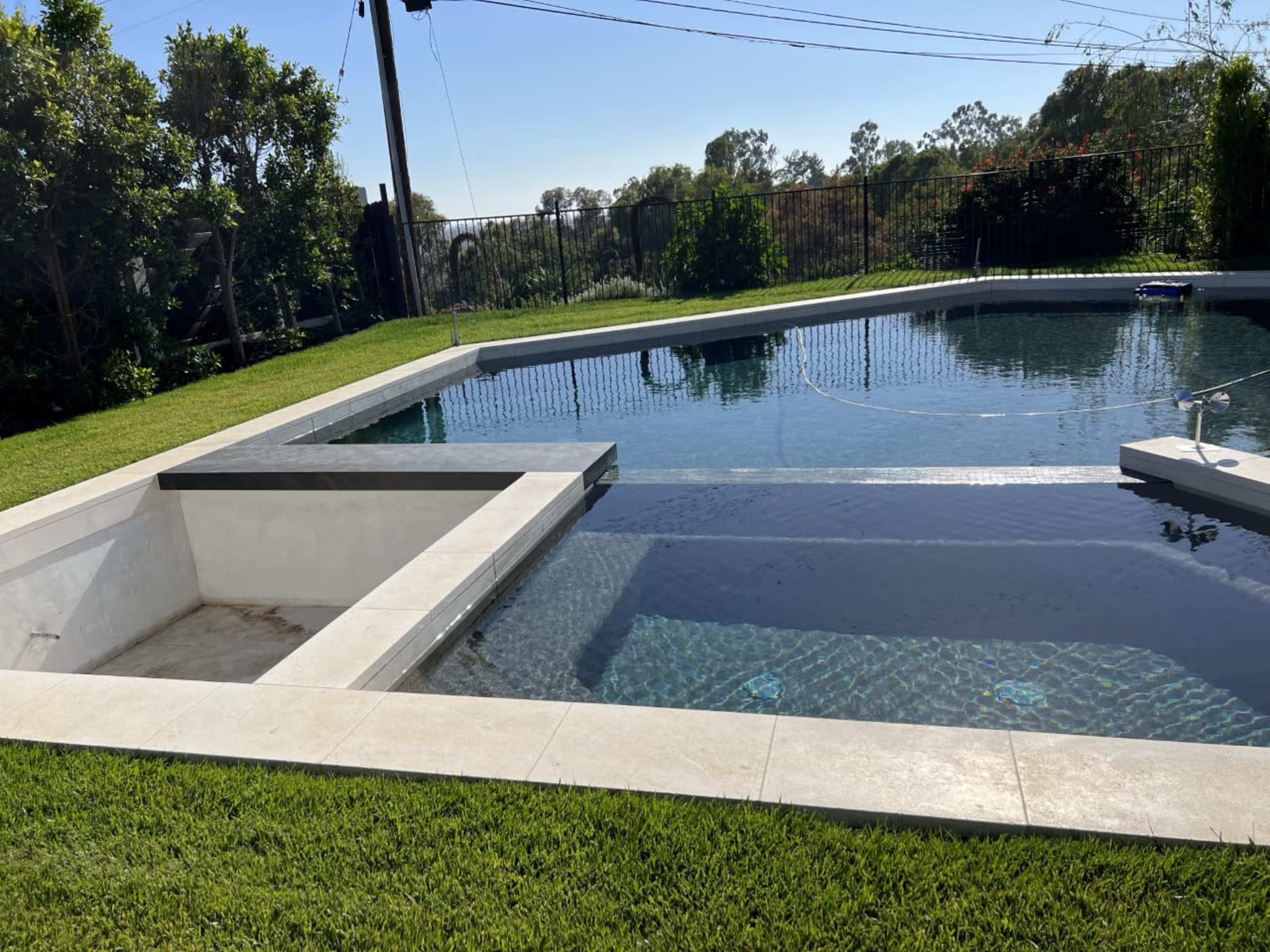 A rectangular swimming pool with a spillover edge and adjacent spa, surrounded by manicured grass and trees.