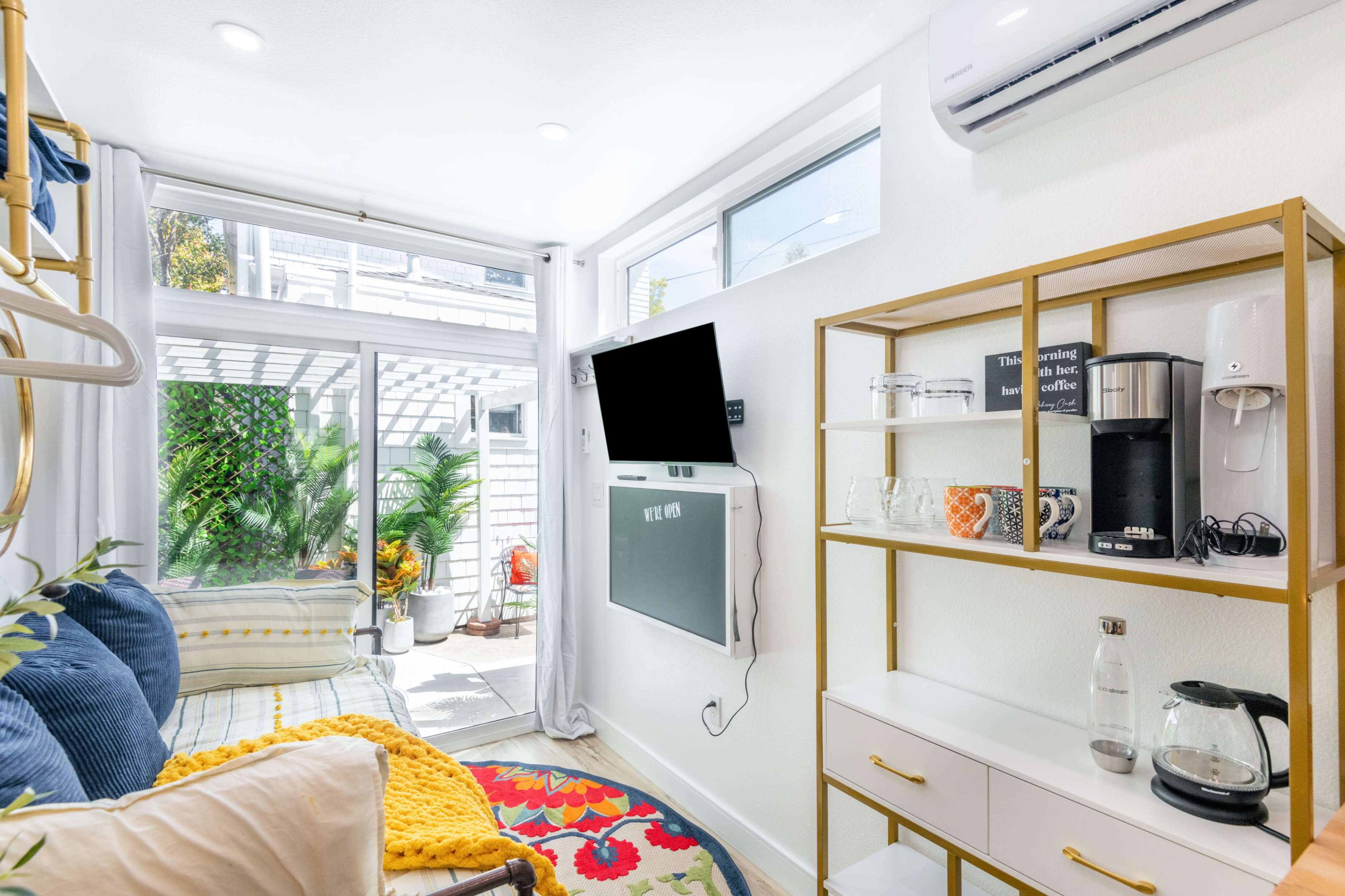 The image shows a bright, modern living space featuring a wall-mounted TV, a shelf with various kitchen appliances, and large windows that offer a view of an outdoor area.