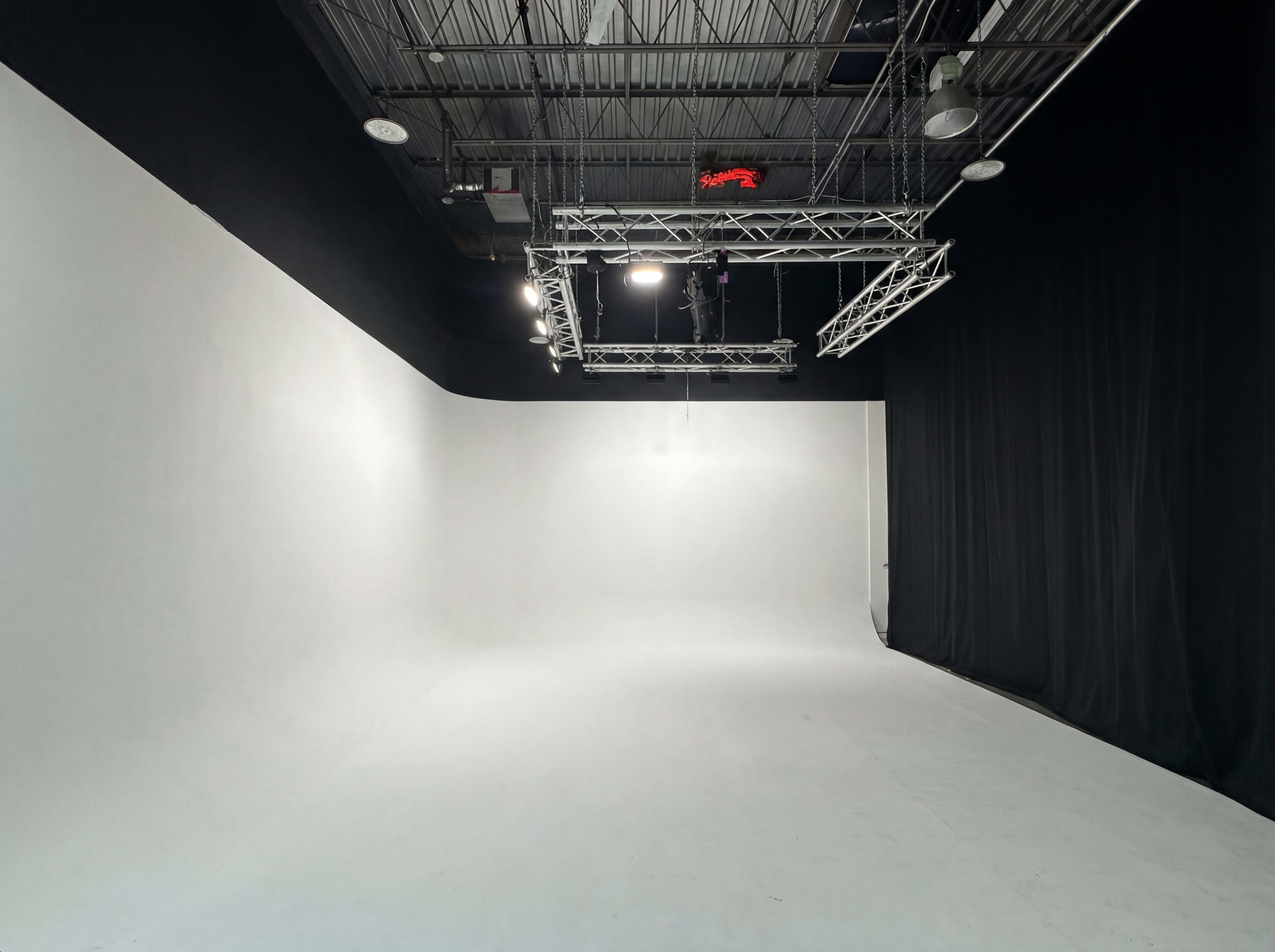 The image shows a spacious empty studio with white walls and a black curtain on one side, illuminated by overhead lights.